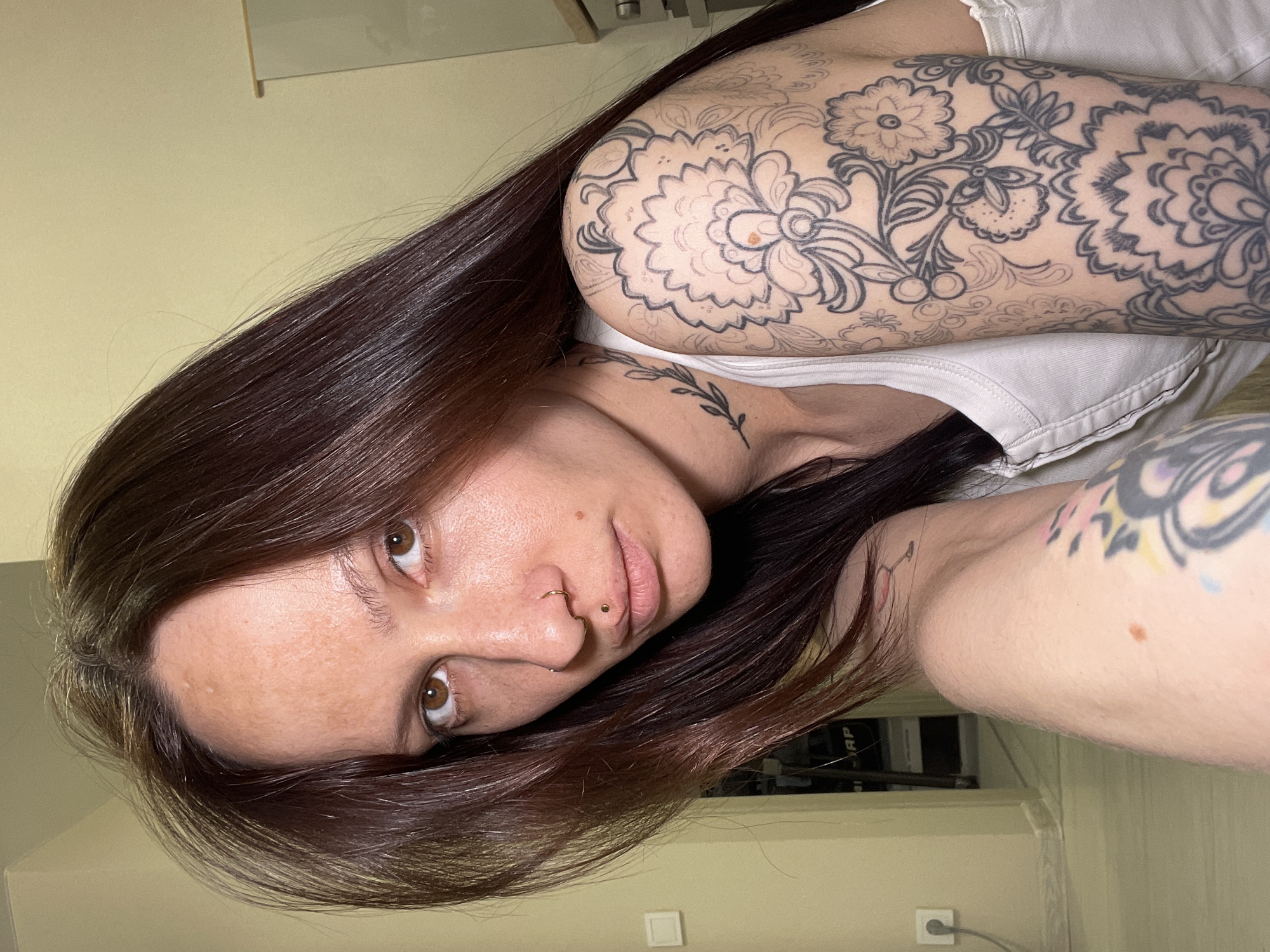A medium shot, rotated 90 degrees clockwise, shows a person lying down indoors in Izhevsk, Russia. The individual, appearing to be female, has long, straight dark brown hair that cascades around their head. They possess fair skin, brown eyes, and a direct gaze towards the viewer. Notable facial features include a gold stud piercing above the upper lip (philtrum piercing) and a small gold hoop in the nose (septum piercing). The skin shows some freckles or moles.
The person is wearing a simple white tank top or camisole. Their right arm (on the left side of the image) is extensively covered with a large, intricate floral tattoo sleeve, featuring designs of flowers and leaves. Parts of this tattoo are fully outlined and shaded with dark ink, while other sections appear to be just the stencil or initial outlines, awaiting completion. A smaller, delicate leaf and vine tattoo is visible on their torso near the armpit. A portion of their left arm (on the right side of the image) is also visible, showing another tattoo that appears to incorporate some pastel colors, such as yellow and light blue.
The background consists of a plain, light yellow or beige wall. In the top left and bottom right corners, elements of the room are visible, including what appears to be a light-colored surface or furniture. A white electrical outlet with a plugged-in cord is visible on the wall in the bottom right, and some dark, indistinct objects, possibly electronic equipment or books, are stacked in the background. The lighting suggests an indoor setting, likely under artificial light.