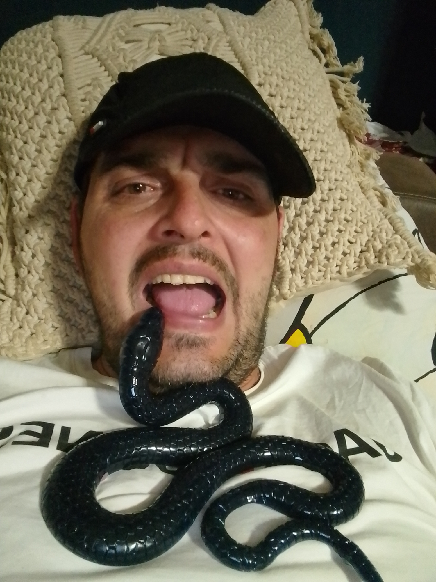This is a selfie-style photograph taken from a close-up perspective, showing a man wearing a black baseball cap and a white t-shirt. He is lying down on a cream-colored, textured blanket or throw. A black, toy snake is placed across his chest and its head is near his open mouth, with its tongue extended. The man's mouth is open, and his tongue is visible. The background appears to be indoors, with a dark wall visible behind the blanket. There is no information available about the specific location in the city of Wedel, Germany.