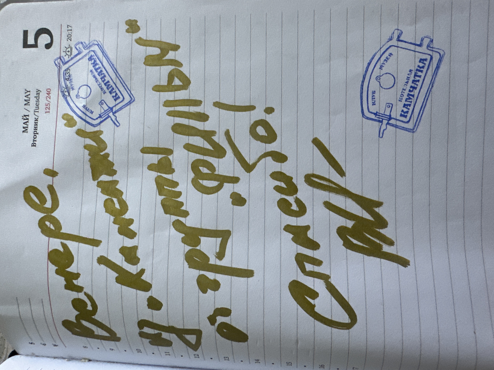The image shows a page in a planner dated May 5th. The day is marked as Tuesday. The page is lined and contains handwritten text in gold ink: "Вечере, J. Karen, гру. "Фильм", Спасибо!". Two blue ink stamps, one on the upper right corner of the page and the other further down, show the words "КЛУБ КОТЕЛЬНАЯ КАМЧАТКА". The stamp's design features a rectangular frame around these words. The stamp's image suggests a possible location connected to the words "КОТЕЛЬНАЯ КАМЧАТКА."