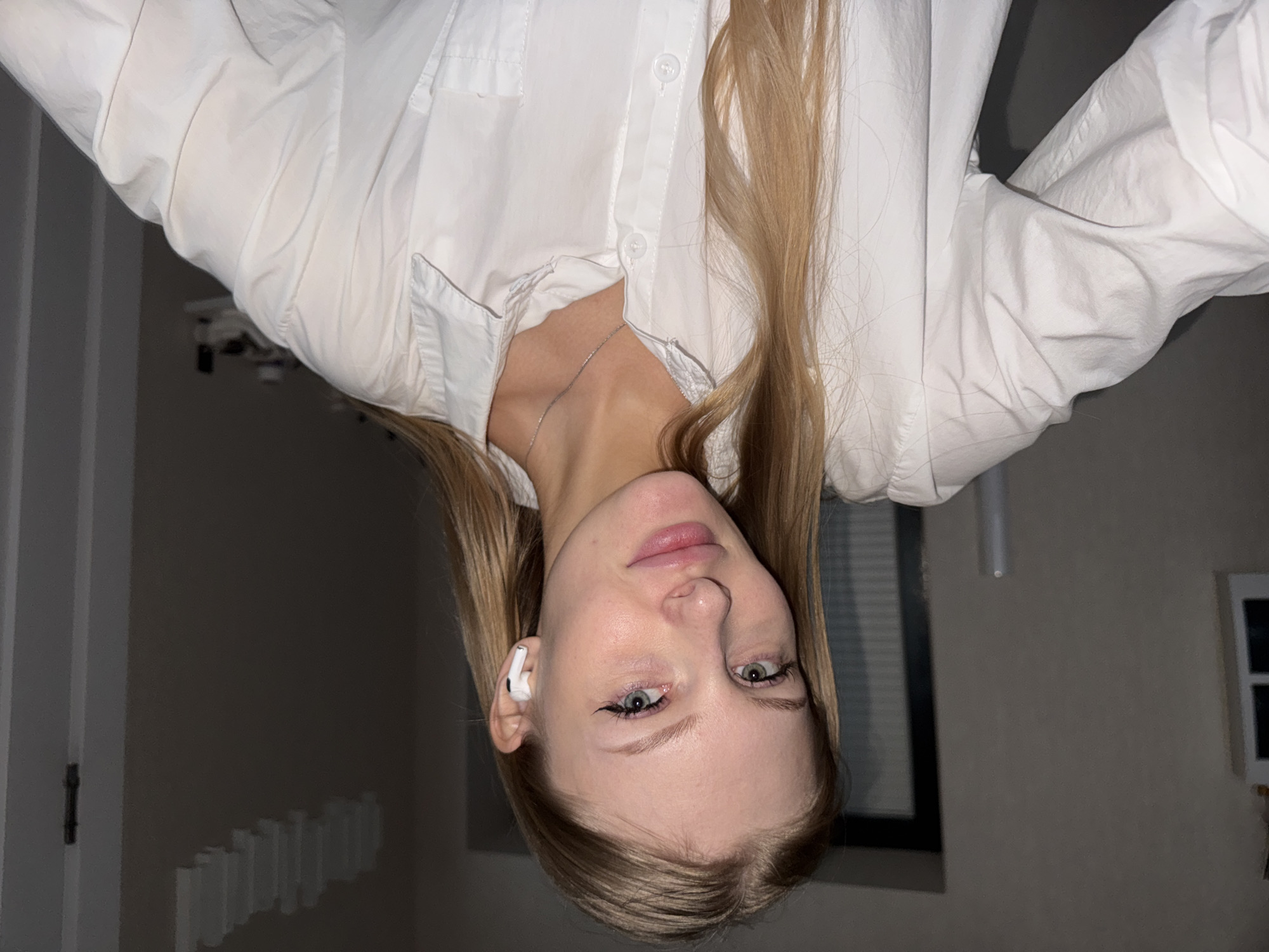 A young woman with blonde hair is shown in an upside-down, close-up shot. She is wearing a white collared shirt and a delicate silver necklace. She has on white earbuds and subtle makeup, including eyeliner and lip gloss. Her eyes are green and her expression appears neutral. The background is dimly lit and includes hints of a room with blinds and a doorway. The image appears to be taken indoors, possibly at night. The location context is Perm, Russia. No text is visible in the image.