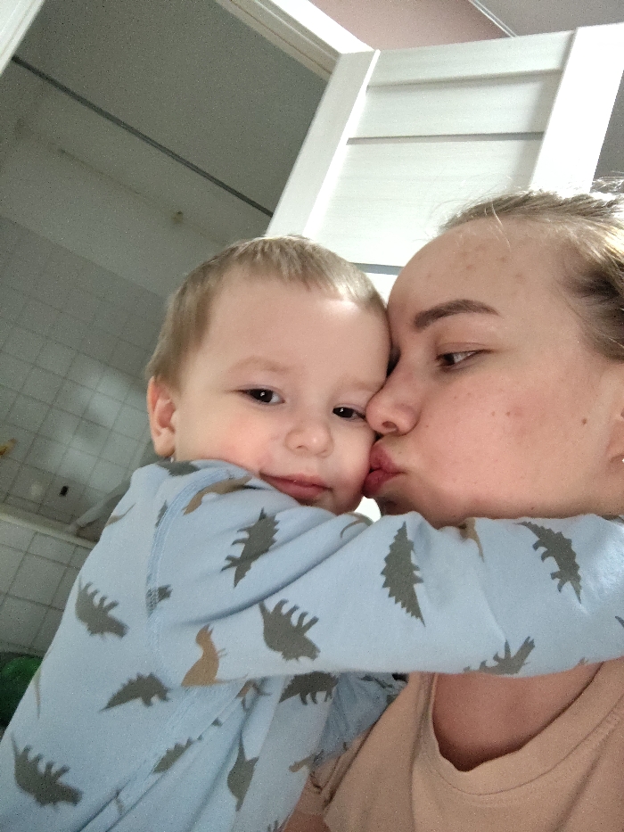A woman with light skin puckers her lips to kiss a child's cheek. The child, wearing a blue dinosaur-print pajama, looks toward the camera, arms wrapped around the woman. The woman wears a tan shirt. Behind them, a white door is visible, and the background suggests a domestic setting with tiled walls. The lighting is soft, highlighting the intimacy of the moment.