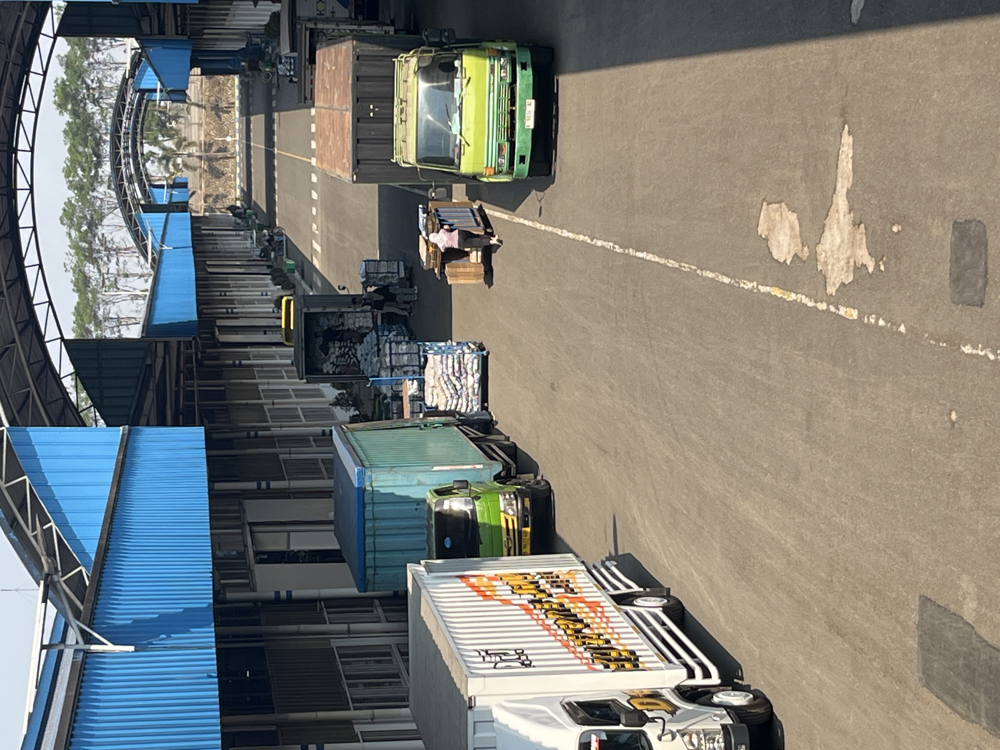 The image depicts an outdoor loading dock or warehouse area in Pelang, Indonesia. Several trucks are present, including a white box truck with orange and yellow decals, a green truck with a blue container, and a green truck parked in front of a large dark gray shipping container. A person wearing a pink shirt and a hijab is pushing a cart loaded with wooden crates and blue bins. Other stacks of goods wrapped in white plastic are visible on the ground. The buildings have blue corrugated metal siding and arched roof structures. The ground is asphalt with white and yellow painted lines. Trees are visible in the background beyond the covered structures. The scene appears to be a logistics or distribution center during daylight hours.