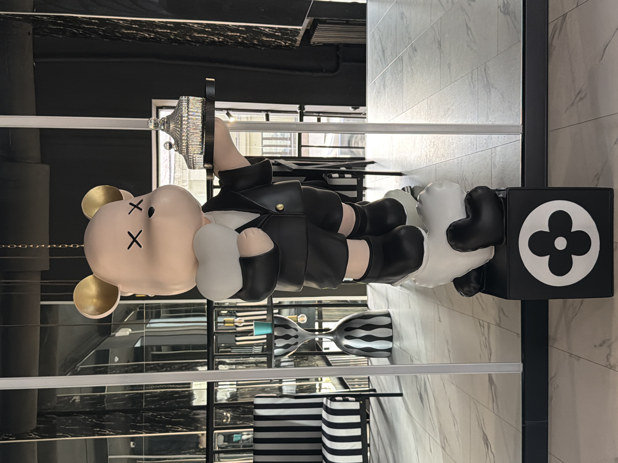 A stylized image depicts a KAWS statue featuring a teddy bear-like figure with crossed-out eyes and wearing a suit. The statue is positioned on a black cube adorned with a white floral emblem. The figure's posture is leaning forward, with white cloud-like appendages serving as hands and feet. The background showcases interior design elements, including striped patterns and decorative objects. The image lacks specifics to determine whether the location is Moscow, Russia.