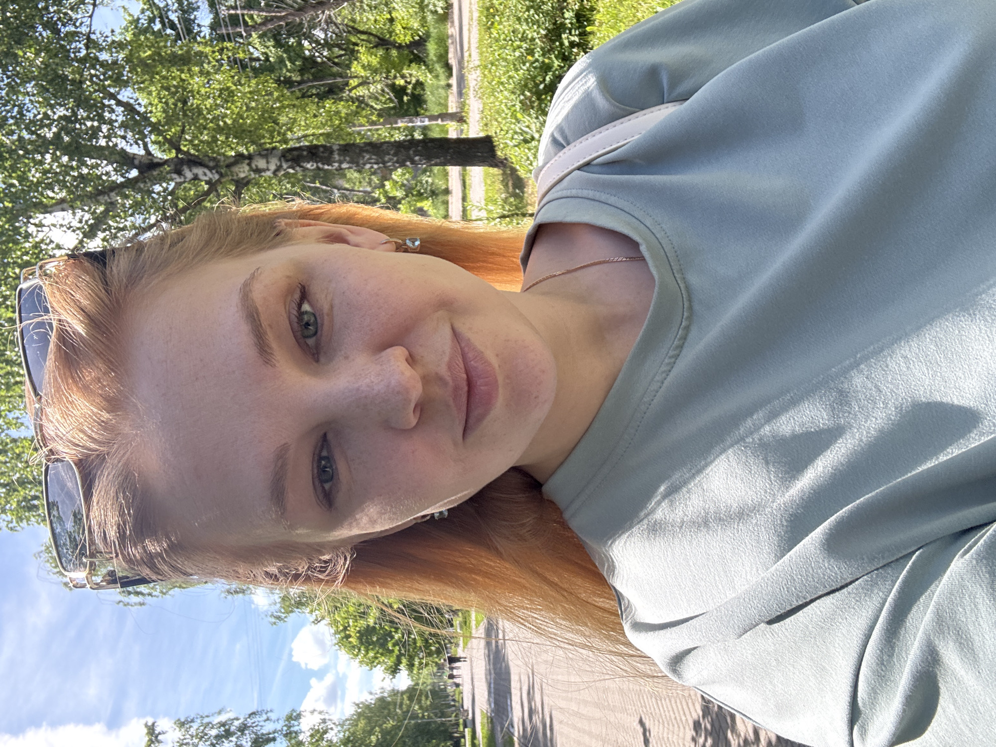 A young woman with reddish-blonde hair, green eyes, and freckles on her face is captured in a bright, outdoor selfie. Her head is tilted slightly, and she looks directly at the camera with a neutral to soft expression. She wears a light teal or mint green long-sleeved top, and a delicate gold-toned chain necklace is visible around her neck. A ring with a light blue stone adorns her right ring finger. Sunglasses are perched on top of her head.
The setting appears to be a sunny day in a park or a green recreational area, possibly in Rusanovo, Russia. The background features lush green foliage from numerous trees, some with distinct pale bark resembling birch. A paved path or road can be seen winding through the greenery further back. The sky is bright blue with scattered white clouds, indicating clear weather. Strong sunlight illuminates the scene, creating defined shadows. No text is visible in the image.