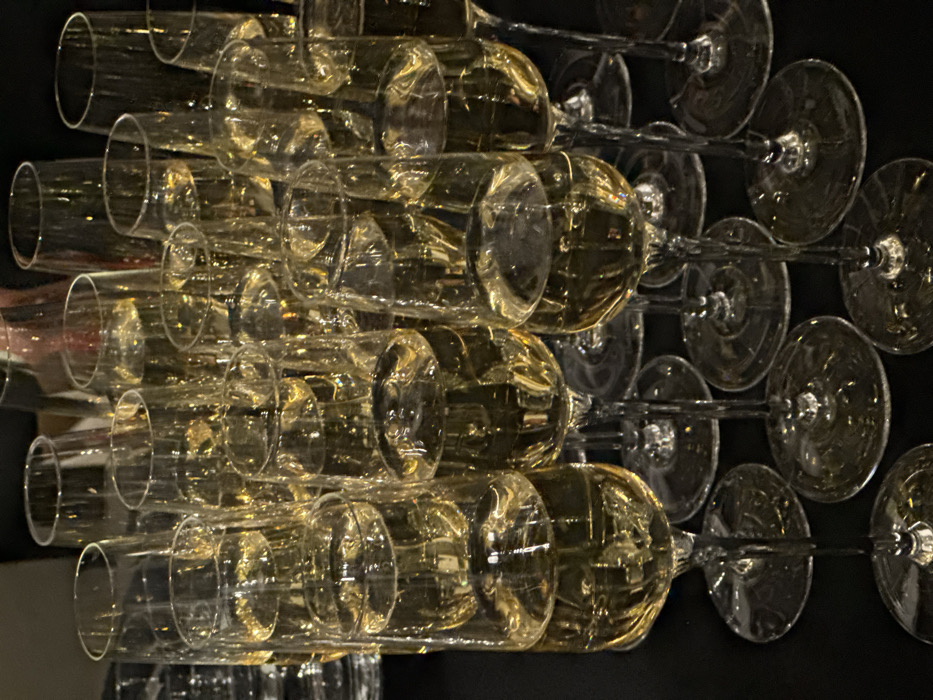 The image depicts a dense arrangement of approximately two dozen clear champagne flutes, each filled with a golden-yellow, effervescent liquid, indicative of sparkling wine or champagne. The glasses feature slender stems and round bases, reflecting ambient warm light from an unseen source. They are positioned closely on a dark, possibly reflective, surface, suggesting preparation for an event. The scene is an indoor setting within the city of Paris, France. No individuals are visible, and no active interactions or specific actions are depicted beyond the static presentation of the beverages.
