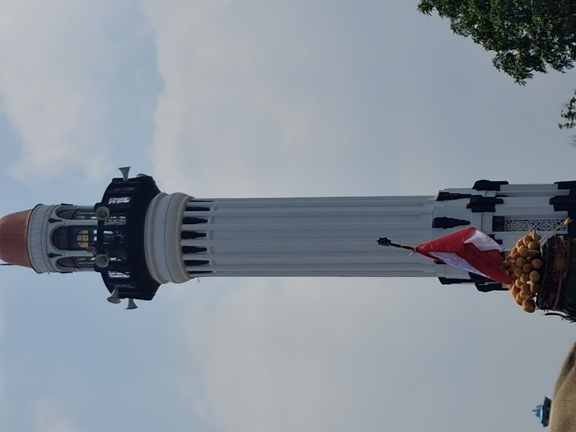 The image shows the top portion of a tall, white minaret with a domed roof that is orange. The minaret has several levels, with balconies featuring decorative black railings and white loudspeakers. The sky is overcast with scattered clouds. In the foreground, at the base of the minaret, there is a display with a stack of round, yellow fruits, a red and white Indonesian flag, and a decorative finial atop a pole. A portion of a green tree is visible on the right side of the image. The minaret is identified as being part of the Great Mosque of Surakarta (Masjid Agung Surakarta) in Surakarta, Indonesia.