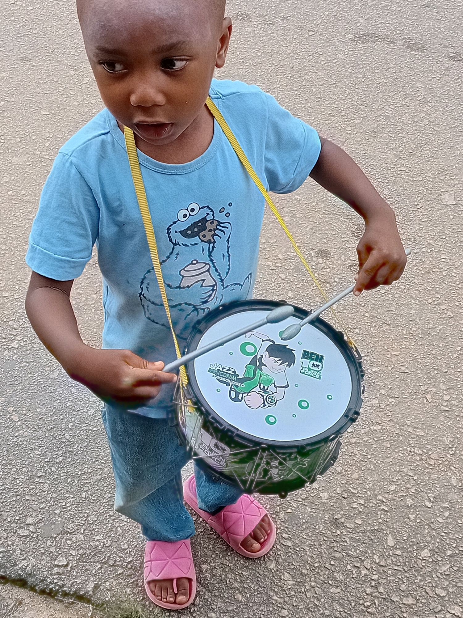 In an outdoor setting on an asphalt surface, likely in Jos, Nigeria, a young Black child stands wearing a light blue t-shirt featuring a Cookie Monster graphic and blue denim jeans. The child holds drumsticks and is playing a drum that hangs from a yellow strap around their neck. The drum has a design of Ben 10 from the animated series "Ben 10: Alien Force." The child is wearing pink slip-on sandals. The background consists of a textured wall and the ground surface.