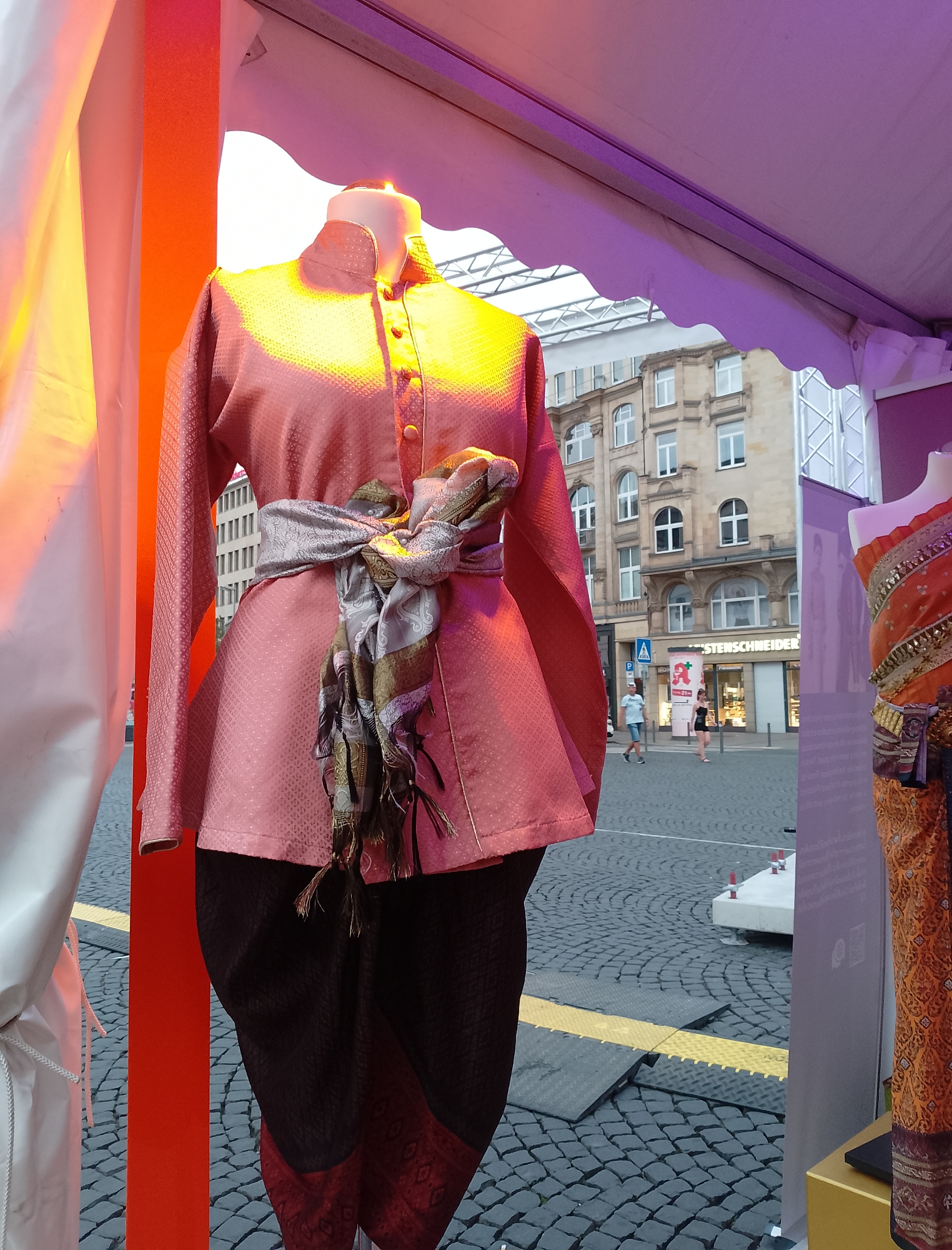 A mannequin in the foreground wears a traditional Thai outfit, consisting of a pink, collared jacket with gold buttons and a patterned fabric, paired with dark trousers featuring a patterned hem. A patterned sash is tied around the mannequin's waist, with tasseled ends. To the right, another mannequin is partially visible, adorned in an orange and gold traditional garment. The mannequins are positioned in what appears to be a city street, paved with cobblestones. In the background, several people are walking past a building with multiple windows, identified by a sign that reads "STERNSCHNEIDER". The scene is illuminated by warm, colored lighting, suggesting an outdoor event or display.