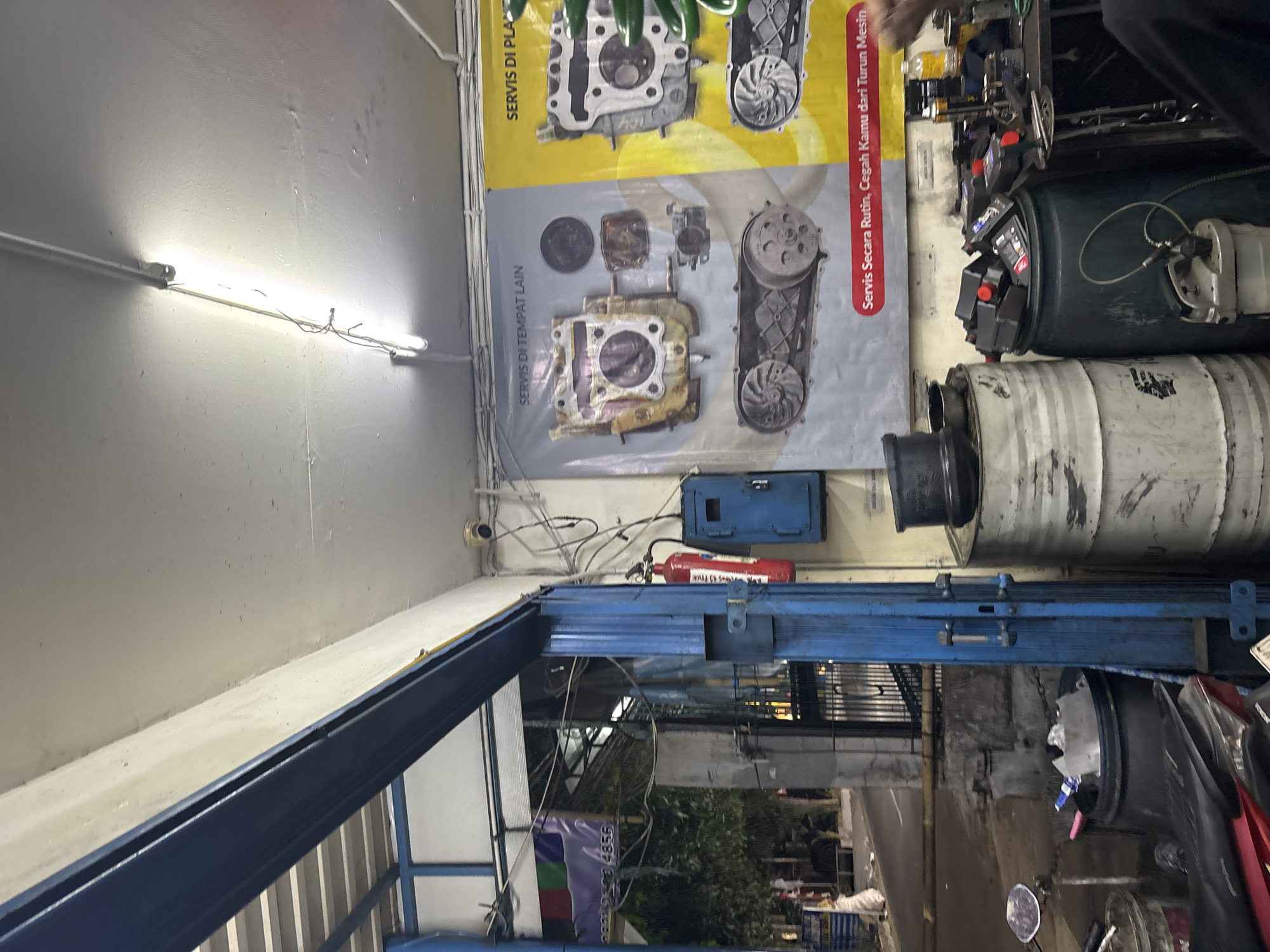 The image depicts the interior of a motorcycle repair shop in North Jakarta, Indonesia, specifically likely in a commercial area given the signage and equipment. The scene is cluttered with various tools, parts, and supplies. Prominently displayed on the wall are two large banners related to motorcycle servicing; one on the left shows engine parts and is labeled "SERVIS DI TEMPAT LAIN" (Service Elsewhere), and another on the right with similar engine parts is labeled "SERVIS DI PLA" (likely Service at Place/Platform). Below these, a red banner reads "Servis Secara Rutin, Cegah Kamu dari Turun Mesin" (Routine Service, Prevent Engine Failure).
In the foreground, on the left side, a red motorcycle is partially visible. Adjacent to it are large, industrial drums, one metallic and others dark green, some filled with what appears to be oil or other fluids. A collection of black oil bottles with red caps is stacked on top of one of the green drums, indicating active use of the shop. A mechanic's workbench with various tools and equipment is visible to the right, with a person's hand and arm in the lower right corner, suggesting ongoing work.
Further back, through an open doorway framed by blue metal structures, a street scene with trees and buildings is visible, suggesting the shop is situated on a street accessible to the public. A red fire extinguisher hangs on the wall near the doorway, mounted next to a blue electrical box. Overhead, fluorescent lights illuminate the space. The overall impression is a busy, functional workshop environment.