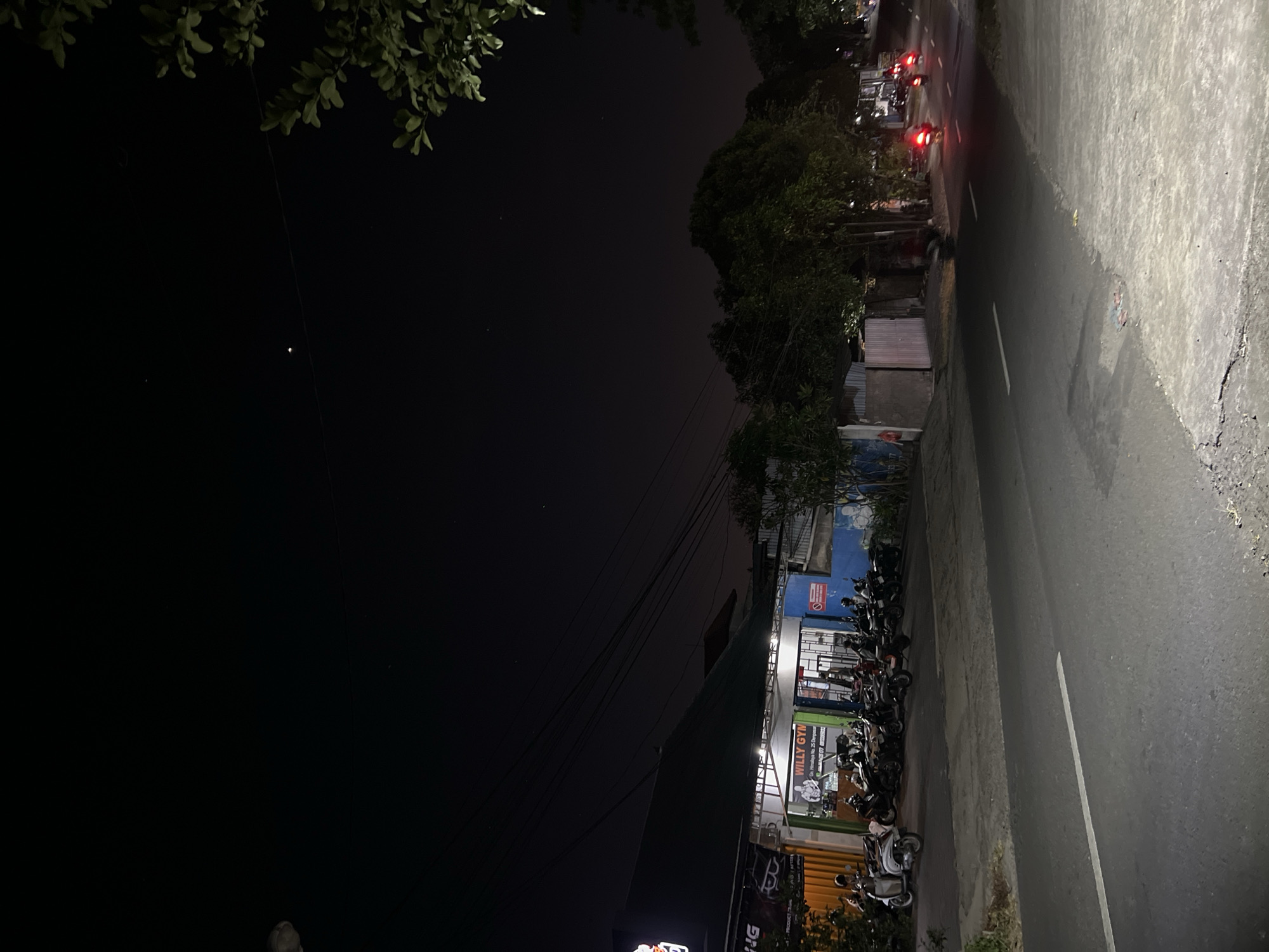 This image depicts a nighttime scene on a street in Denpasar, Indonesia, likely in the vicinity of a commercial area. In the foreground, a paved road with a white dividing line runs from left to right. To the left of the road, a building with an awning is illuminated by interior lights. Signs are visible, including one that reads "Willy Gym" with an image of a muscular figure. A row of parked motorcycles, mostly scooters, is lined up in front of the building. Some people are visible near the motorcycles and in front of the gym.
Beyond the building, a line of trees forms a dense green barrier. In the mid-ground, the road continues and appears to have at least three motorcycles in motion, indicated by their red taillights, suggesting they are moving away from the viewer. The sky above is dark with a few faint stars visible. A bright object, possibly a planet or a distant light, is prominent in the upper central portion of the frame. The overall impression is a typical street scene at night in an urban or suburban Indonesian setting.