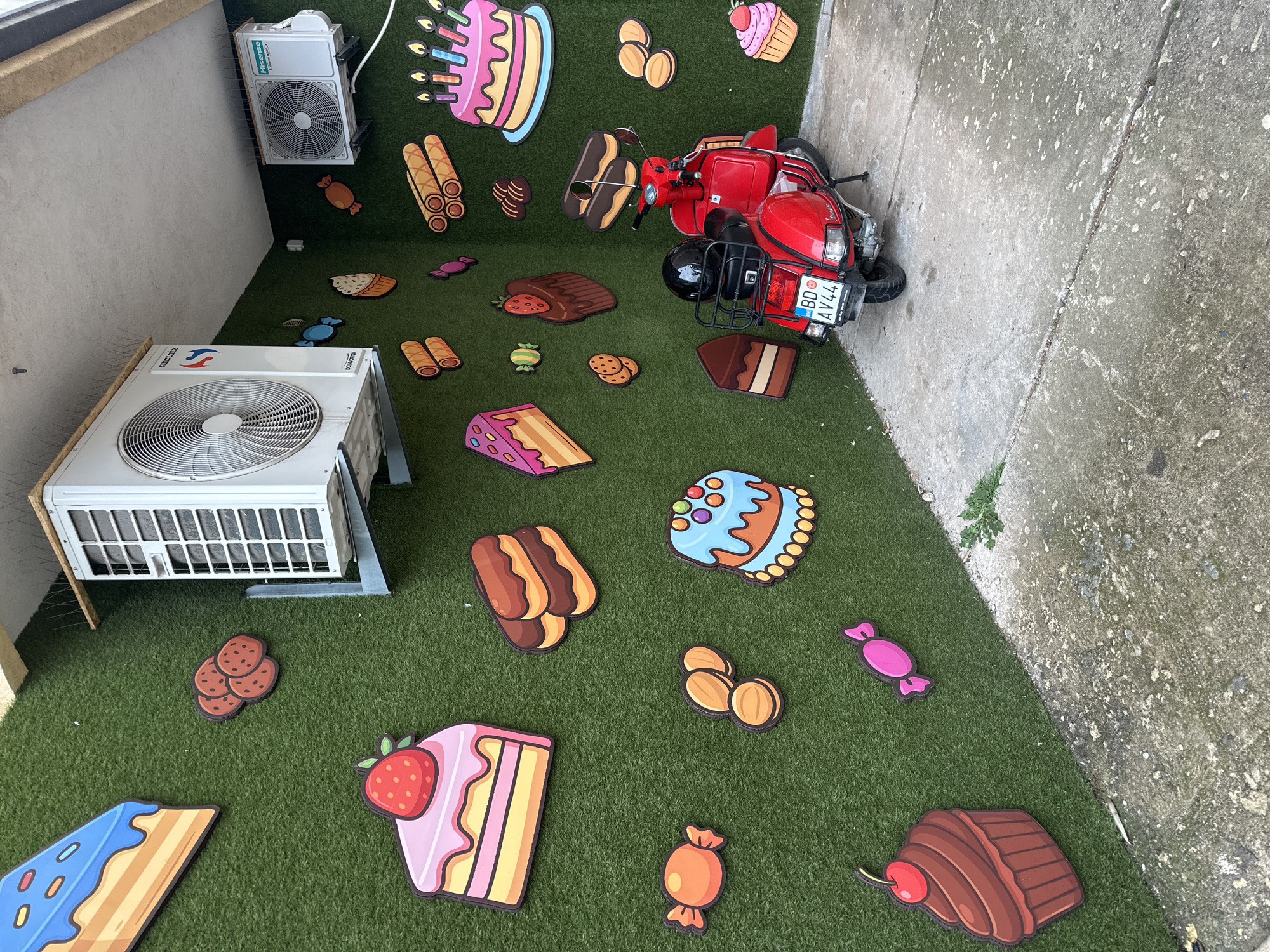A red Vespa-style scooter is parked in the corner of an outdoor area in Budva, Montenegro. The scooter displays a license plate, "BD AV44," and a black motorcycle helmet rests on its seat, below a black rear luggage rack. The two visible walls of the corner are covered with artificial green turf, which is decorated with numerous cartoon-style cutouts of desserts and candies, including various cakes, cupcakes, cookies, and wrapped sweets. Two white outdoor air conditioning units are mounted on these walls; one "SINCLAIR" unit on the left and one "Hisense" unit on the right, both equipped with circular fan grilles. Bird deterrent spikes are present along the ceiling edge above the air conditioning units. The ground consists of a discolored, cracked concrete slab with sparse green vegetation emerging from the cracks and along the base of the walls. No individuals are present in the scene.