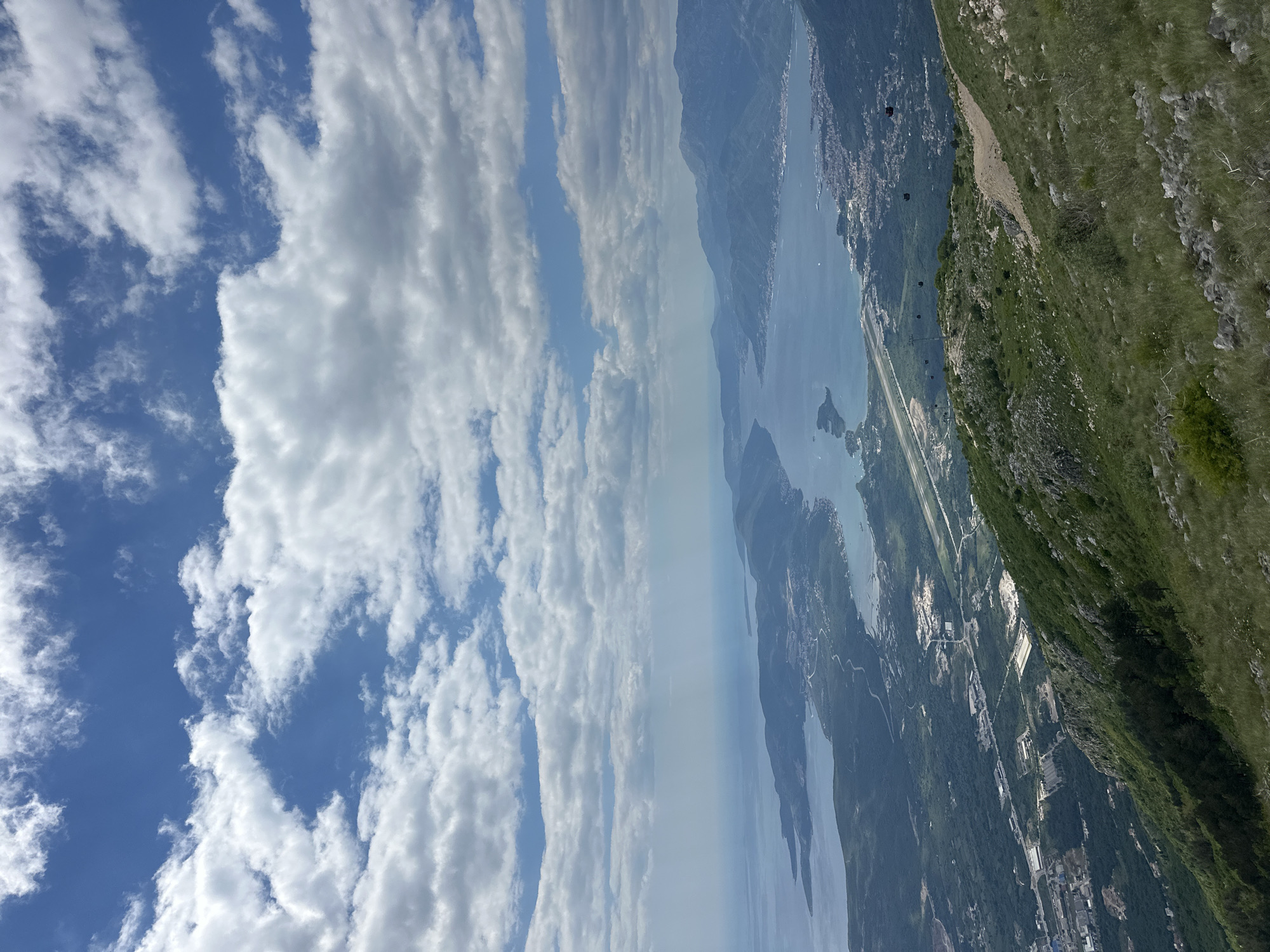 The image shows a landscape in Radanovići, Montenegro. The sky features alternating bands of blue and white clouds. A long, narrow body of water extends into the distance, surrounded by mountains. Several towns or settlements are visible along the coastline. A green, grassy slope occupies the lower-right portion of the image. Cable cars are visible above the landscape.