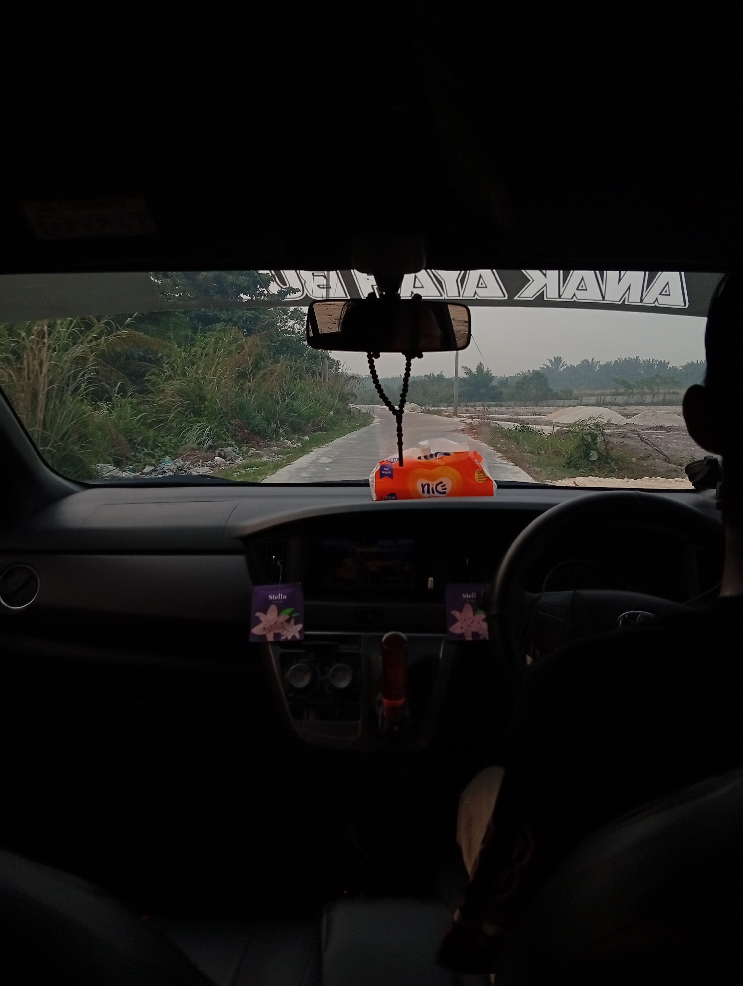 The image depicts the interior view from the driver's seat of a vehicle, looking out through the windshield. A road stretches into the distance, flanked by lush greenery and some construction materials. The dashboard is visible, with a pack of "Nice" brand tissues placed on it, alongside a beaded car air freshener hanging from the rearview mirror. A "Stella" air freshener is also visible hanging from the dashboard. The steering wheel and part of the driver's head are in the foreground, suggesting the photo was taken by the driver. The weather appears overcast, with a hazy sky in the background. The location is identified as Teluk Kenidai, Indonesia.