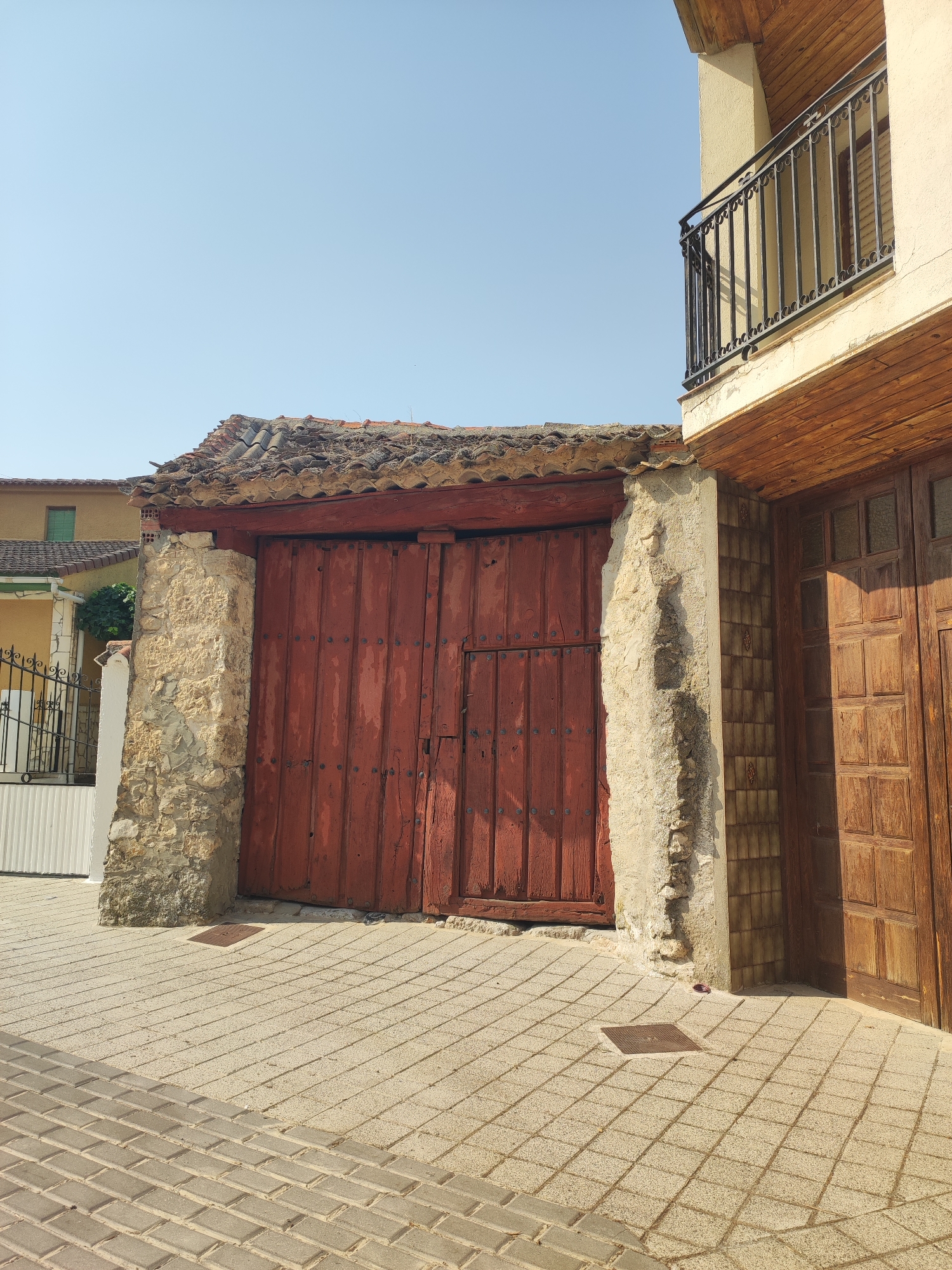 The image shows a street in Traspinedo, Spain, with a large, weathered red wooden door set into a stone entryway. The door is studded with metal bolts and appears to be old. Above the door is a rustic tile roof. To the right of the red door, there is a section of wall covered in small, dark rectangular tiles, and beyond that, another large wooden door. A balcony with a black metal railing is visible on an upper story of the building to the right. The ground in front of the doors is paved with light-colored rectangular tiles. The sky above is clear and blue. To the left of the stone entryway, a white fence with metal railings is visible, as well as a portion of a yellow building.
