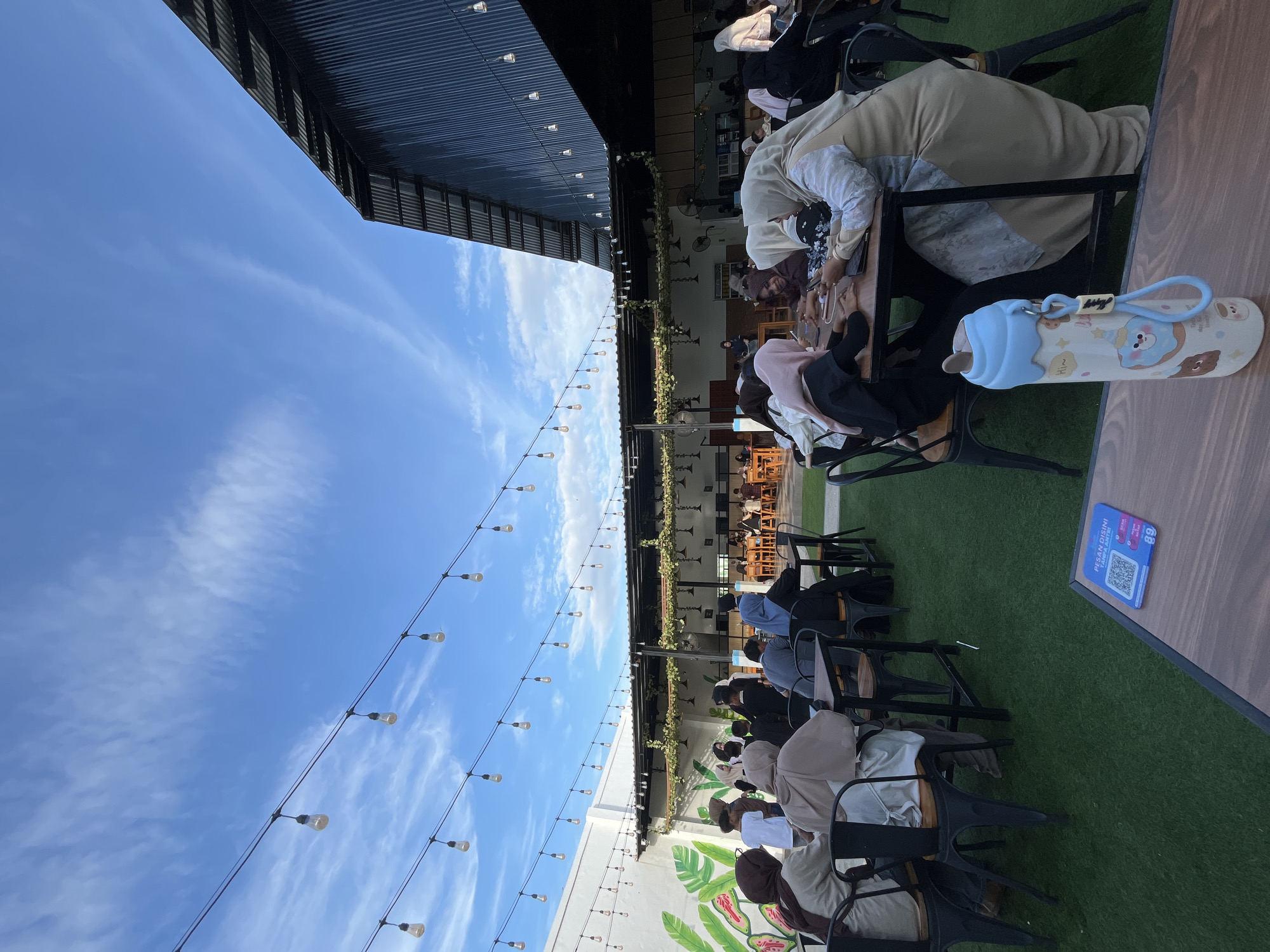 This image depicts an outdoor dining or cafe area in Lhokseumawe, Indonesia, under a bright blue sky adorned with scattered wispy white clouds. Numerous string lights with visible clear bulbs are suspended overhead, crisscrossing the scene. The ground of the dining area is covered in green artificial turf, upon which dark-colored tables and chairs are arranged.
To the left, a plain white wall features a large, stylized mural of green and pink leaves. To the right, a substantial building with a dark, corrugated metal roof and wall is visible, slanting sharply upwards. In the mid-ground, a semi-covered structure, likely part of the cafe, is adorned with hanging greenery and additional pendant lights beneath its roof. Beyond this structure, more tables and individuals are present in a further covered section.
The scene is populated by a diverse group of individuals, predominantly adults. Many women are observed wearing headscarves (hijabs). In the immediate foreground on the right, several women are seated at dark wooden tables. One woman in a beige hijab and a light-patterned dress is partially visible, looking down, possibly at a mobile device. Another woman in a dark top and hijab is similarly engaged, as is a woman in a light-pinkish hijab with her back mostly to the camera. On the left foreground, a woman in a dark brown hijab and another in a light beige hijab are seated with their backs to the viewer. Across the area, people are engaged in conversation, using mobile phones or tablets, and dining.
On the table closest to the camera, a white water bottle with a light blue top, a cartoon bear design, and "Hi~" text is prominently displayed. Adjacent to it is a blue rectangular card with a QR code and Indonesian text that reads "PESAN DISINI SAMBIL ENTRI" (Order here while entering), along with the number "89," indicating a digital menu or ordering system.