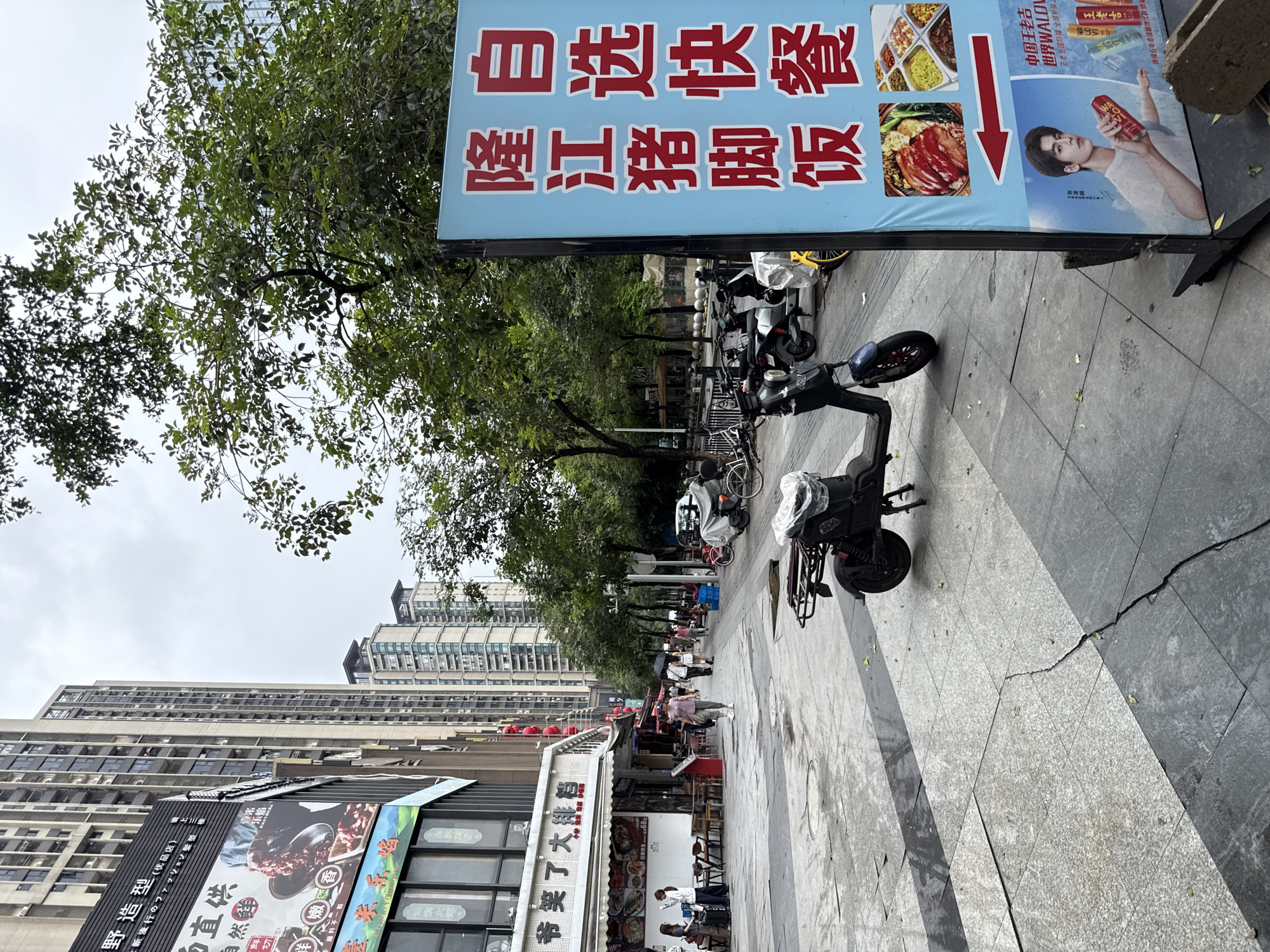 The image depicts a street scene in Haizhu District, China. On the right side of the frame, a tall, light blue sign with red Chinese characters reads "自选快餐" (Self-service fast food) and "隆江猪脚饭" (Longjiang Pig Trotter Rice). Below the text are images of food dishes, a red arrow pointing left, and a poster of a young man holding a can of "WALOVO" beverage. The sign is placed on a sidewalk, adjacent to a street lined with tall buildings. In the foreground, a dark blue electric scooter is parked. The street is paved with stone tiles, some of which are wet and reflective. In the background, several other buildings, trees, and people walking are visible. Some individuals are carrying umbrellas, suggesting a possibility of recent rain or cloudy weather. The buildings appear to be modern high-rises. The environment suggests a commercial area with shops and eateries.