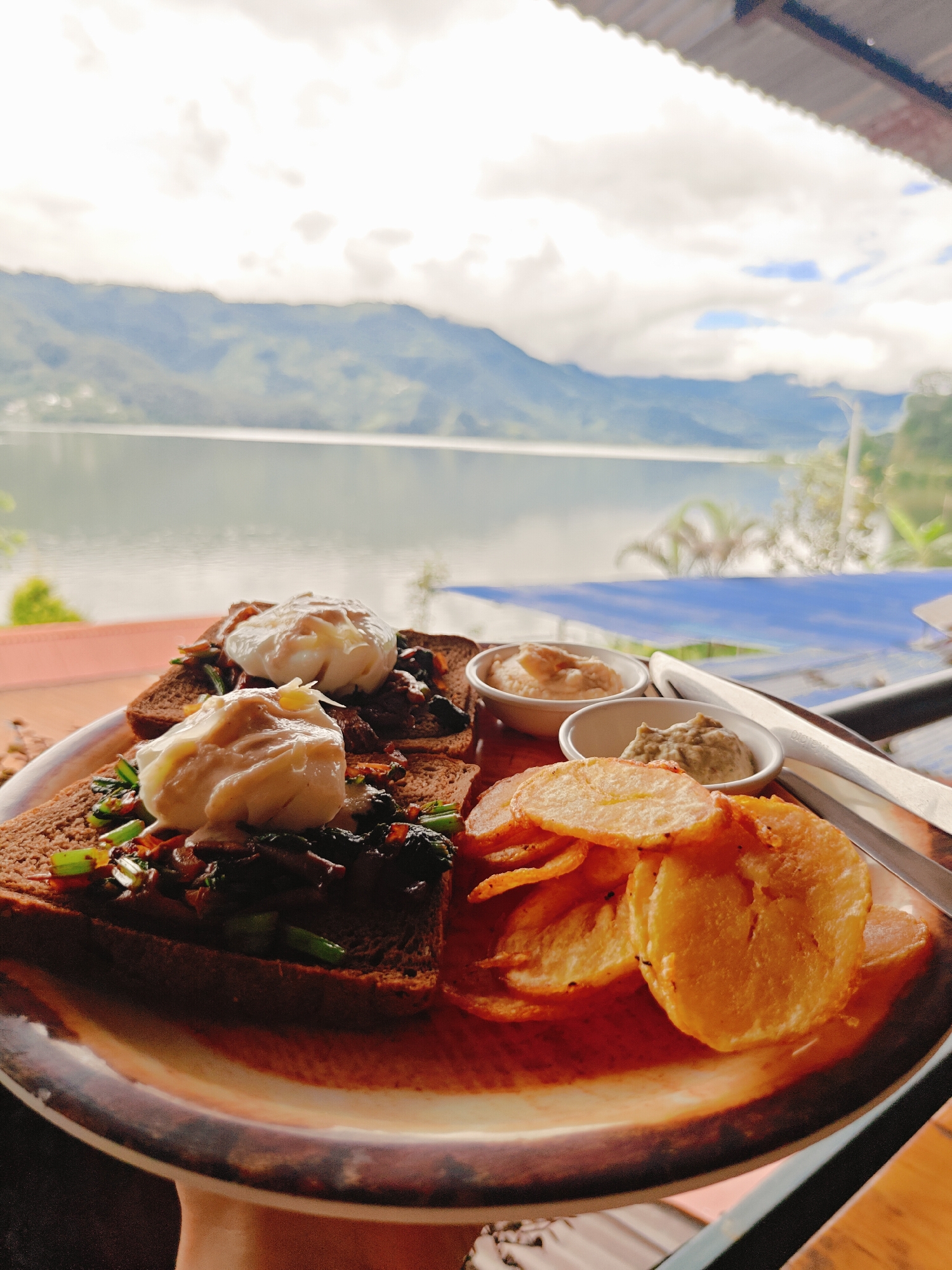 A close-up, eye-level shot captures a plate of food with a scenic mountain and lake view in Pokhara, Nepal. The plate, with a textured brown rim, contains two pieces of toast topped with poached eggs and chopped vegetables. To the right, a pile of golden-brown potato chips sits beside two small white bowls filled with dips. In the background, a vast lake stretches towards a misty mountain range under a cloudy sky. The overall scene suggests a meal enjoyed with a picturesque vista.