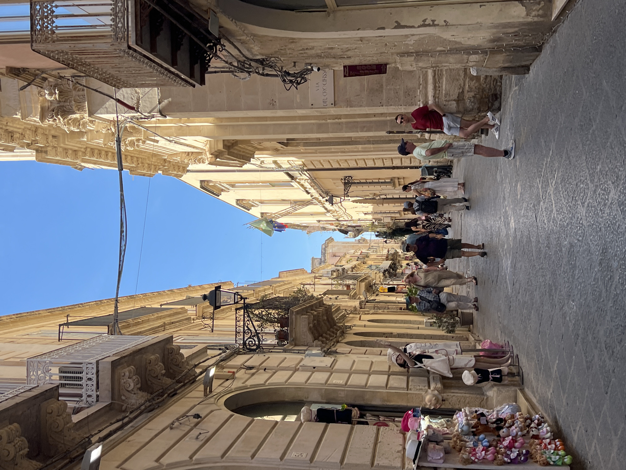 The image depicts a narrow, pedestrianized street in the historic center of Syracuse, Italy, likely Ortigia. Tall, cream-colored buildings with ornate balconies and architectural details line both sides of the street. A bright blue sky is visible above. The street is paved with dark cobblestones.
Several people are walking and standing in the street. On the left side, a shop displays an array of stuffed animals and hats. A female mannequin dressed in a hat and apron stands outside the shop. Further down the street, a group of people are walking away from the camera. On the right side, two men are conversing near a building facade. One man in a red t-shirt and grey shorts leans against the wall. Another man in a light green t-shirt and striped shorts stands opposite him. A street sign that reads "VIA DEL CROCIFISSO" is visible on the right-hand building. A surveillance camera is mounted near the sign. Wires are strung across the street, connecting buildings and streetlights. The overall scene conveys a sense of a warm, sunny day in a charming, old European city.