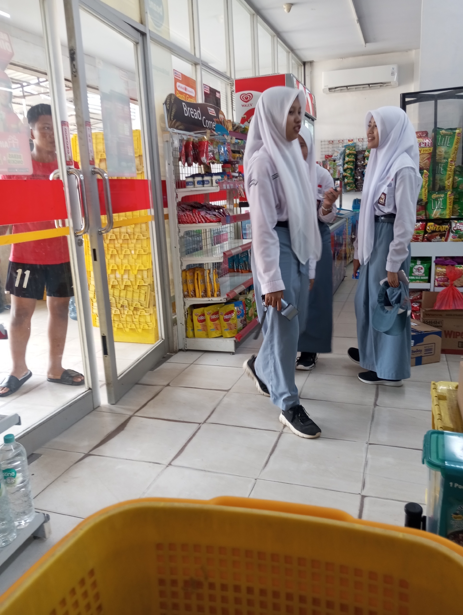 In a convenience store in Tanjung Gading, Indonesia, a young man wearing a red shirt and black shorts with the number "11" visible stands inside the entrance, looking towards the left. Two young women in white hijabs and school uniforms are walking away from the entrance, towards the interior of the store. The woman on the right is carrying a blue cap in her hand, and the woman on the left is holding what appears to be a phone or small object. Shelves stocked with various snacks and drinks are visible behind them. The floor is tiled, and a yellow shopping basket is partially visible in the foreground. The lighting suggests an indoor environment, possibly during daytime.