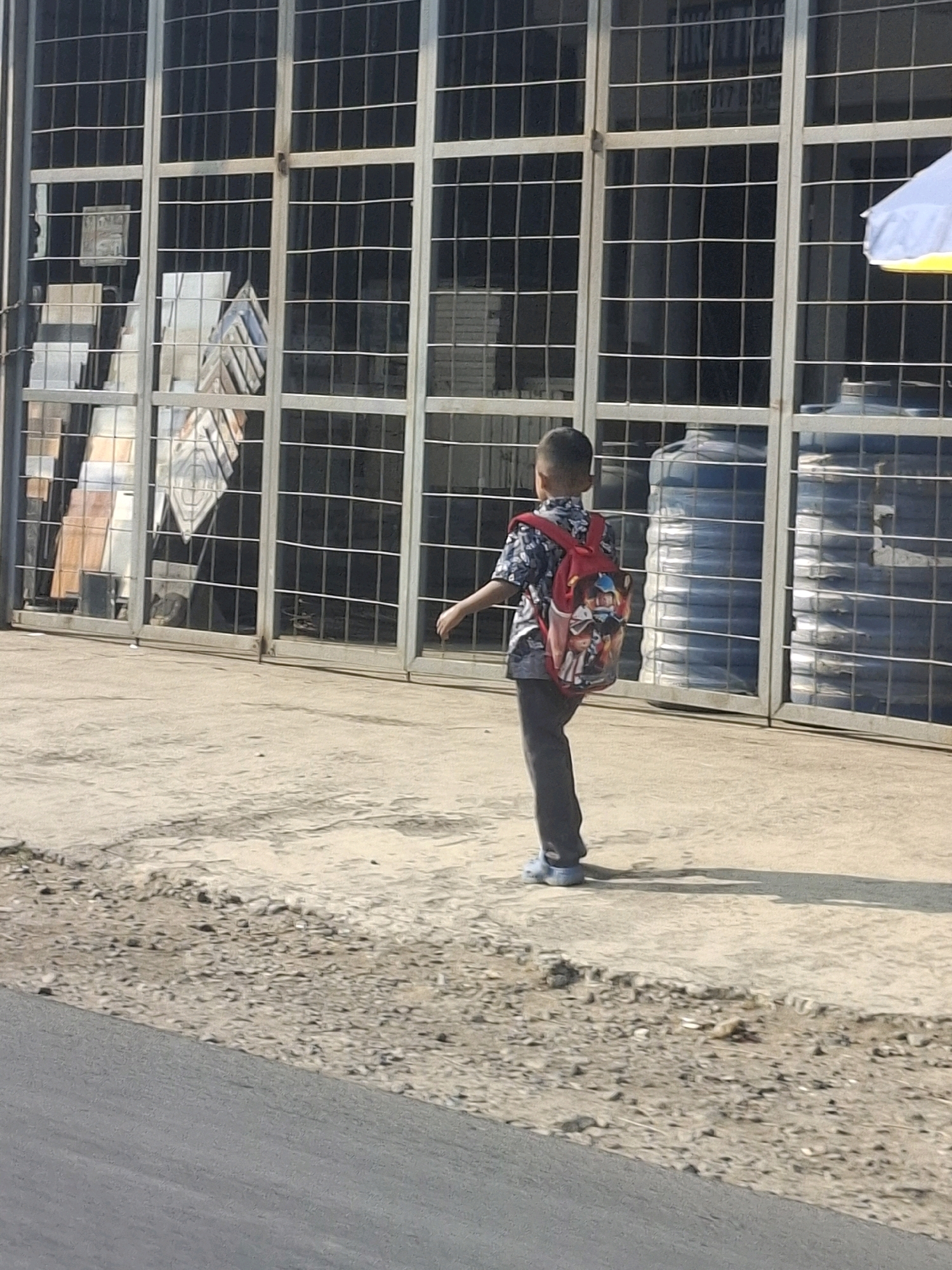 A young boy wearing a patterned shirt, dark pants, and blue shoes with a red backpack is walking away from the camera on a concrete sidewalk. The backpack has cartoon characters on it. Behind the boy is a metal grate structure with various items visible through it, including what appear to be tiles and large blue water containers. The scene is lit by sunlight, casting shadows on the ground. The environment suggests an outdoor urban setting.