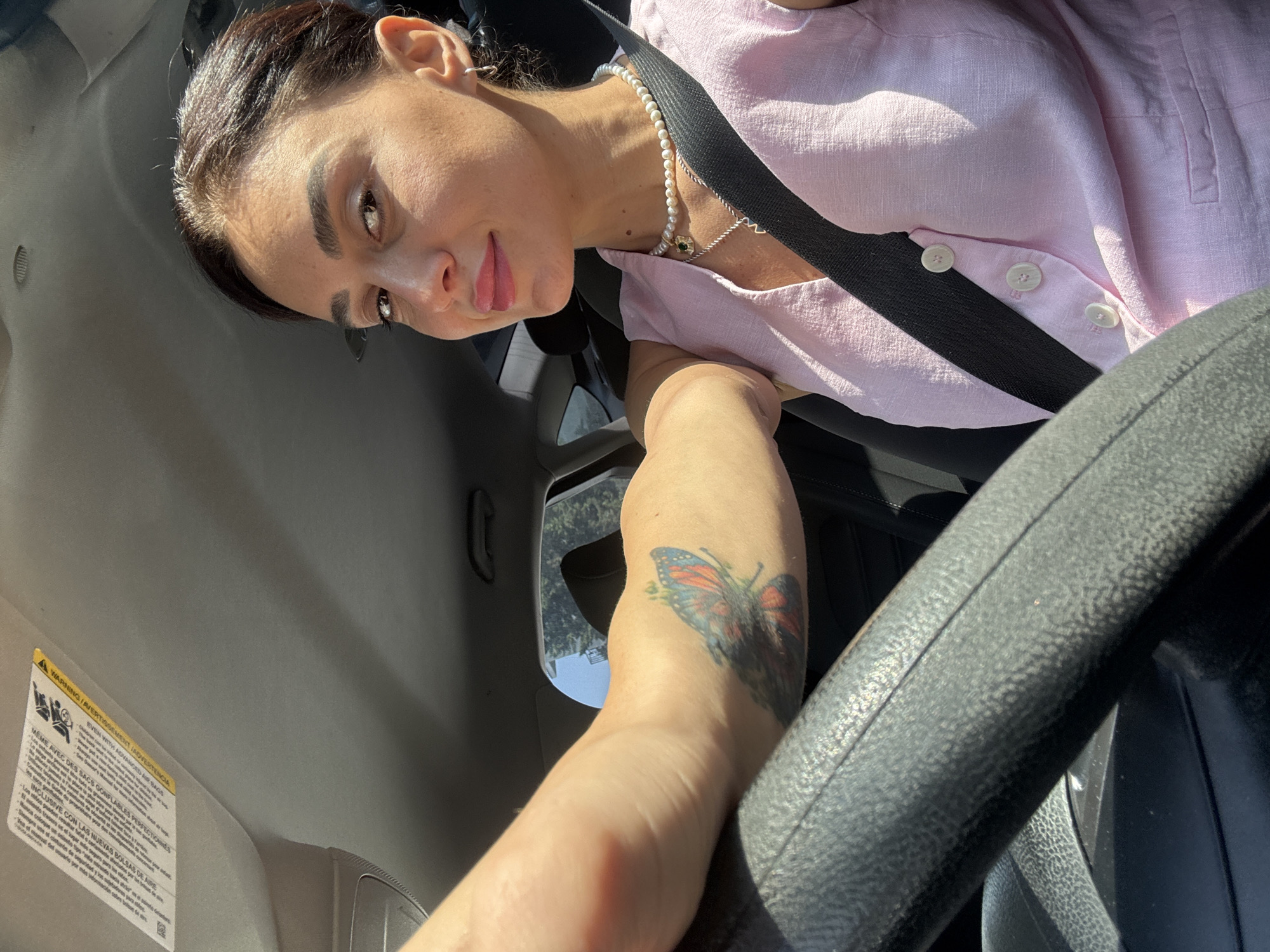 An overhead interior shot captures a woman with dark hair pulled back, wearing a pink sleeveless top and a pearl necklace, seated in the driver's seat of a vehicle. Sunlight streams in from the right, illuminating her face and upper body. Her left arm rests on the steering wheel, revealing a colorful butterfly tattoo. The car's interior, including a sun visor and seatbelt, is visible. The scene is set during the daytime in Tbilisi, Georgia, as indicated by the car's sun visor which features warning labels in multiple languages, including Georgian.