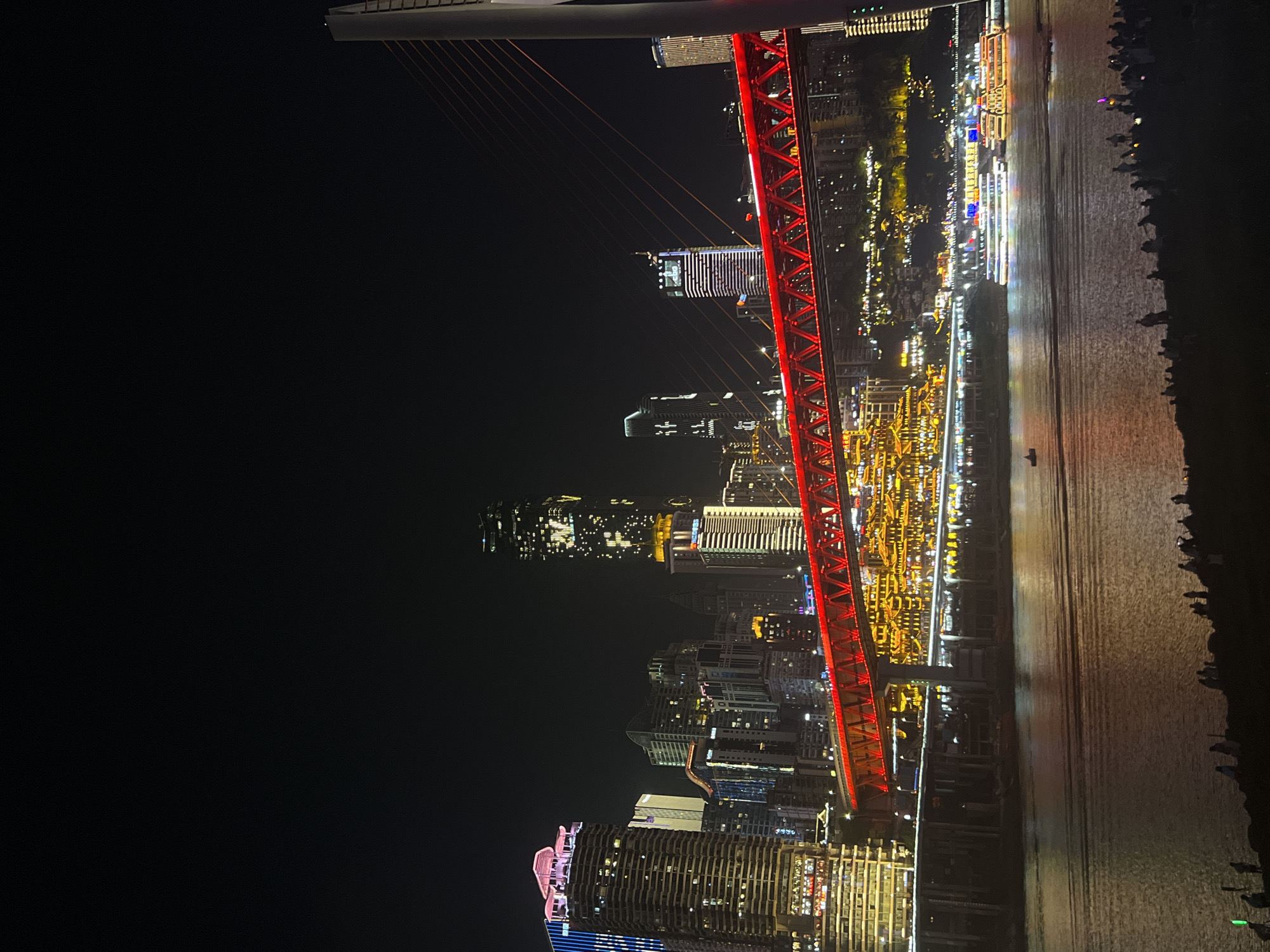 This is a nighttime, eye-level, full shot of a modern cityscape in Jiangbei Cheng Street, China, viewed from across a wide body of water. The prominent feature is a vibrant red suspension bridge spanning the water, illuminated with numerous lights. Behind the bridge, a dense collection of illuminated skyscrapers of varying heights and architectural styles dominate the skyline. Lights are visible in many windows, and some buildings are adorned with colorful exterior lighting. Several tour boats with their own lights are cruising on the water. Along the foreground water's edge, silhouetted figures of people are gathered, seemingly enjoying the nighttime view. The sky is dark and clear, providing a stark contrast to the bright city lights.
