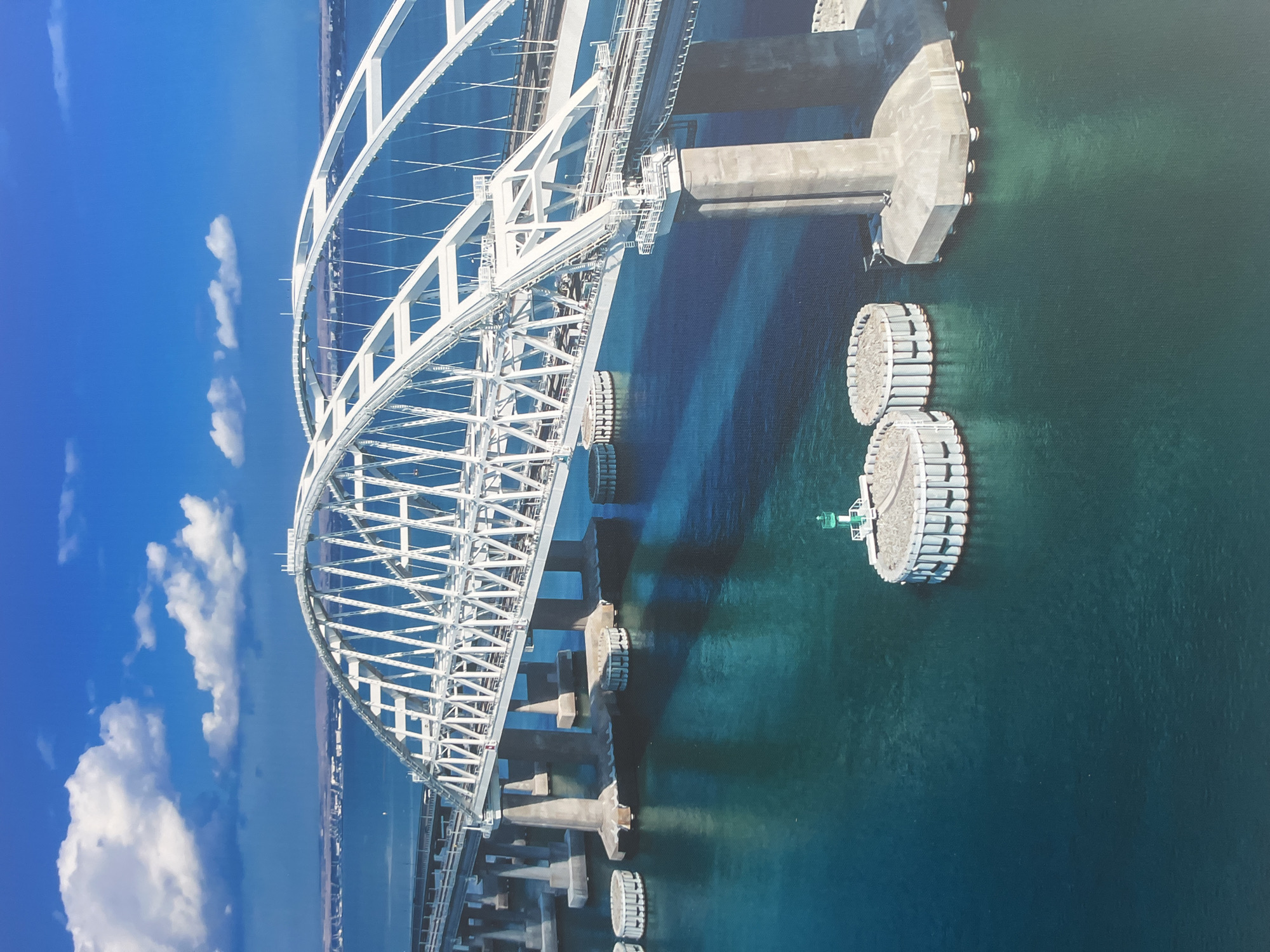 The image depicts the Kerch Strait Bridge, a massive infrastructure project connecting Russia's Taman Peninsula to the Kerch Peninsula of Crimea. The bridge is an impressive feat of engineering, featuring multiple spans and arches. It crosses a body of water that appears to be calm and a deep blue-green color. In the distance, a shoreline with some structures can be seen under a bright blue sky with scattered white clouds. The bridge itself is a prominent white structure, with its intricate metal framework visible. Several concrete piers support the bridge above the water. In the foreground, circular structures are visible in the water, likely navigational aids or support elements for the bridge. One of these circular structures has a green beacon on top. The scene suggests a clear day with good visibility, highlighting the grandeur and scale of the bridge against the natural backdrop of the sea and sky. The city of Минзаг is not depicted in this image.