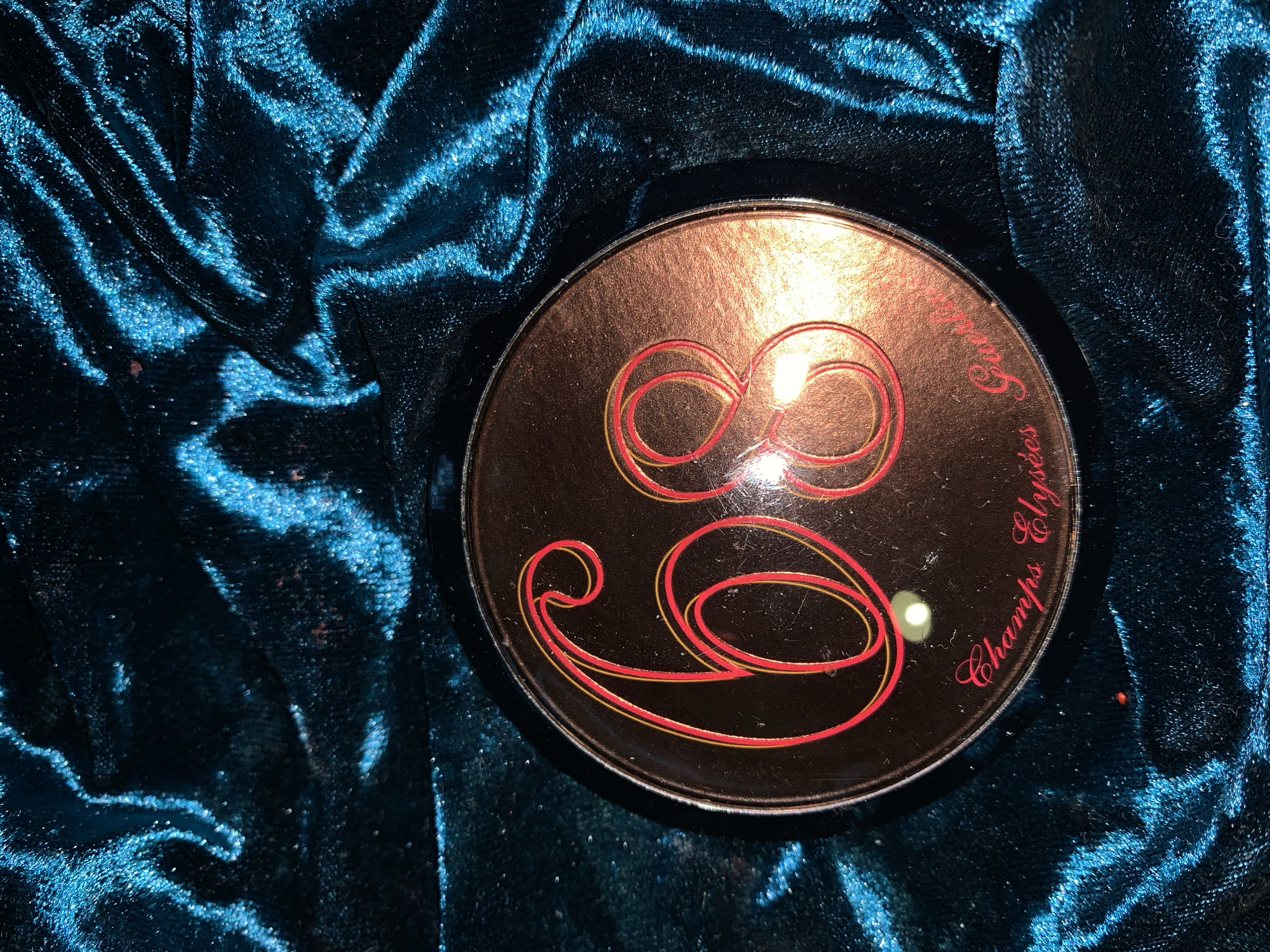 A close-up photograph captures a round, reflective gold-toned compact placed on a backdrop of folded, shimmering blue velvet fabric. The compact's surface is adorned with red stylized figures resembling cursive numerals and the inscription "Champs Elysées".