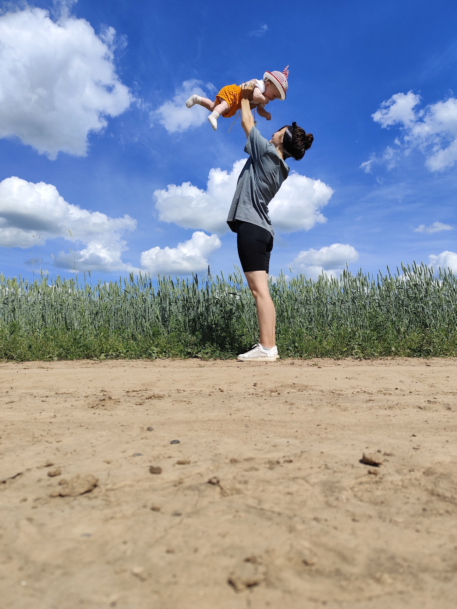 A person is tossing a baby into the air against a bright blue sky with fluffy white clouds. The person is wearing a grey t-shirt, black shorts, and white sneakers. They have their hair tied up in a bun and are looking up at the baby. The baby is wearing an orange outfit with white polka dots and a red and white striped hat. The baby's legs are extended in the air.
The scene takes place outdoors, possibly in a rural area. A field of tall green grass or wheat is visible in the background, behind the person and baby. A dirt path runs in the foreground, suggesting a rural road or trail. The lighting indicates it is daytime, likely with sunny weather given the clear blue sky and bright clouds. The overall mood is playful and joyful, captured by the action of the person tossing the baby. The location is specified as Фоки, Russia. There is no visible text in the image.