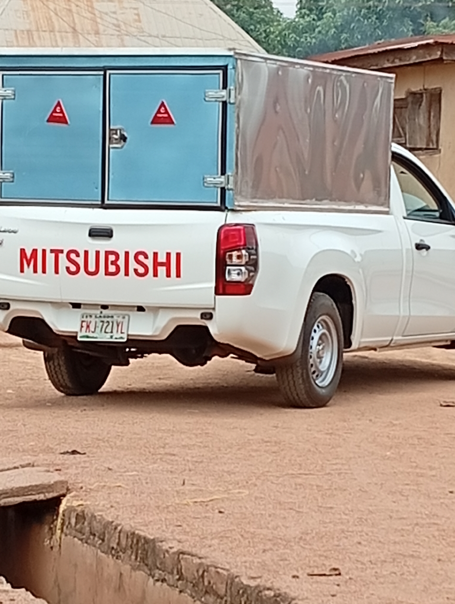A white Mitsubishi pickup truck is parked outdoors, viewed from its rear three-quarter angle. The truck features a custom-built cargo box in its bed, which is light blue at the back with two doors, and metallic silver on its visible side. Each blue door has a red triangular warning sign with a white symbol, possibly an 'A', inscribed within it. The tailgate of the truck clearly displays the word "MITSUBISHI" in prominent red lettering.
A Nigerian license plate, partially visible, reads "FXJ-721YL". Above the numbers, "BENUE STATE" is faintly discernible, consistent with the provided location context of Gboko. The vehicle's rear wheel, a standard steel rim, is visible, resting on unpaved, reddish-brown ground.
In the immediate foreground, a concrete and stone open drainage channel or gutter runs across the image. The background is somewhat blurred, showing simple structures, possibly houses with corrugated roofs, and green foliage under what appears to be an overcast sky or during the late afternoon, given the soft lighting. No specific activity is depicted; the truck appears stationary. No people are visible. The scene suggests a setting in a rural or semi-urban environment in Gboko, Nigeria.