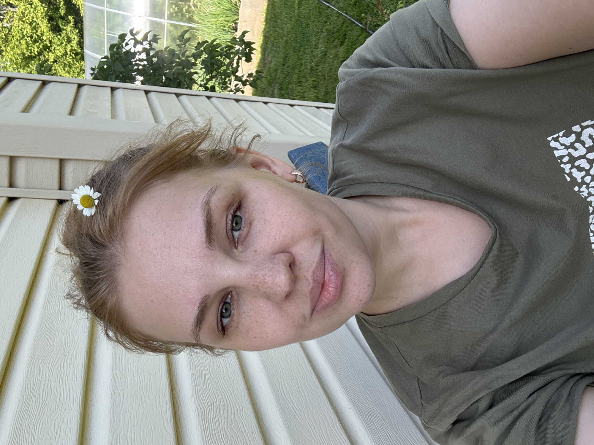 A young woman with light skin and freckles, light brown hair tied up with a daisy clipped in the back, is taking a selfie outdoors. She is wearing a plain olive green t-shirt. She has green eyes and is looking directly at the camera. Behind her, a light-colored textured wall is visible, possibly the side of a house. To the right, a section of a greenhouse and green foliage are present. The background suggests a suburban or rural setting. The woman appears to be in a relaxed, natural pose. The location is the village of Dunilovo, Russia.