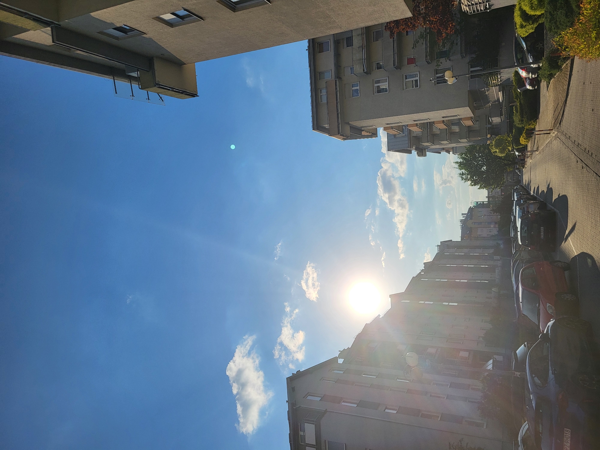 The image shows a street scene in an urban area, likely in the city of Unknown, Unknown. The view is looking upwards towards the sky. Two buildings frame the top left and top right of the image, with the sky visible between them. The sky is blue with some scattered white clouds. The sun is shining brightly, positioned towards the middle-right of the frame. A street runs along the right side of the image, with parked cars lining the side of the road. The buildings are multi-story apartment blocks with windows and balconies. Trees and greenery are interspersed along the street and around the buildings. There are no people visible in the image.