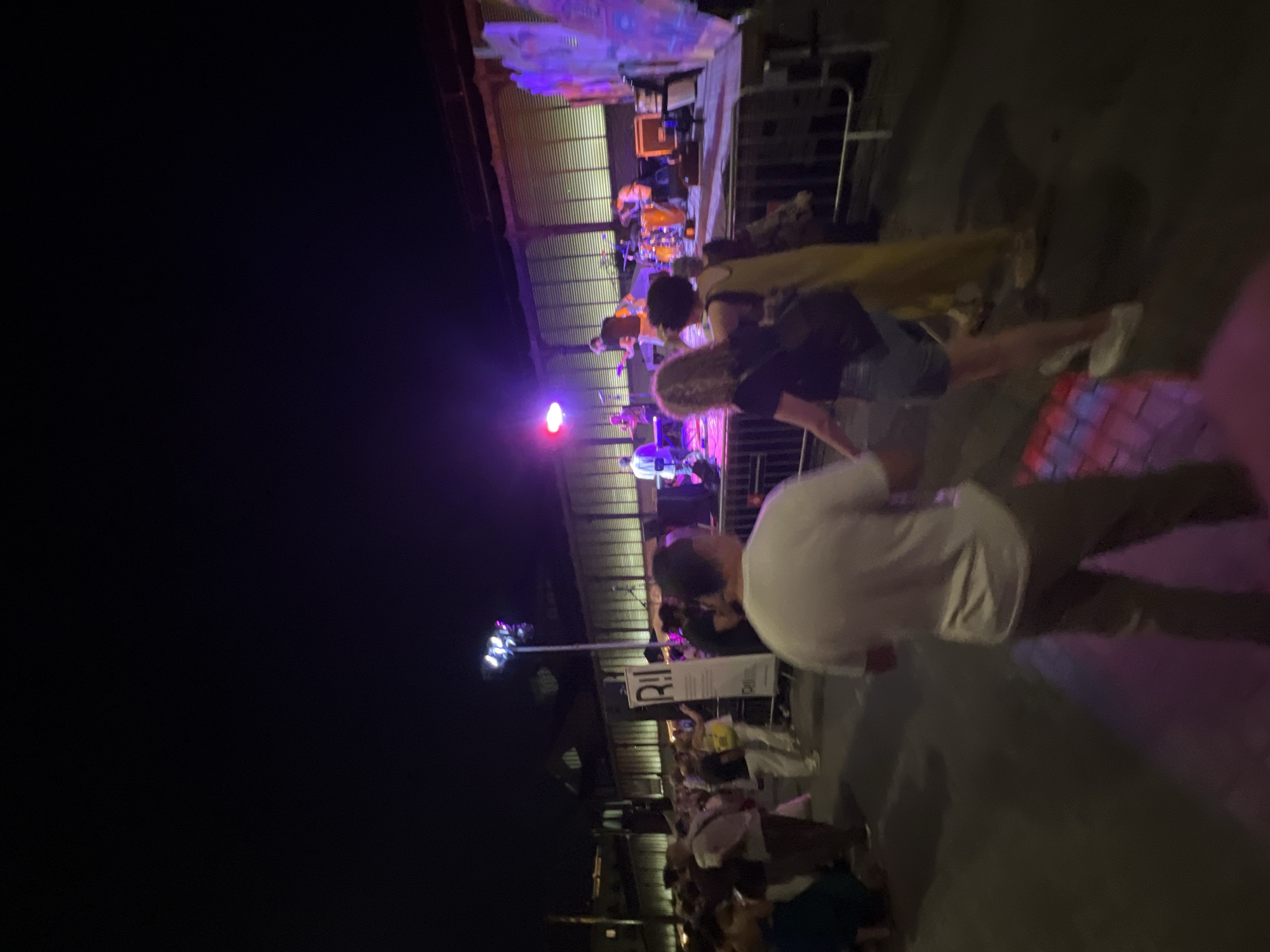 The image shows a nighttime street scene in Barcelona, Spain, likely during an outdoor event or festival. In the foreground, a person in a white t-shirt and khaki pants walks away from the camera, their back turned. To their right, a woman with curly blonde hair wearing a black t-shirt and denim shorts is visible, holding hands with another person whose face is obscured. Behind them, a stage is set up with musicians performing under purple and pink lights. Several other people are visible in the midground, some gathered near the stage and others walking along the street. A tall banner with the "R!!" logo is displayed on the left side of the image, suggesting a branded event. The background features a long, striped building, possibly a market or industrial structure, with lights illuminating its facade. The ground is asphalt, with patches of colorful light reflecting on it, indicating the presence of lighting effects. The overall atmosphere is one of a lively outdoor gathering at night.