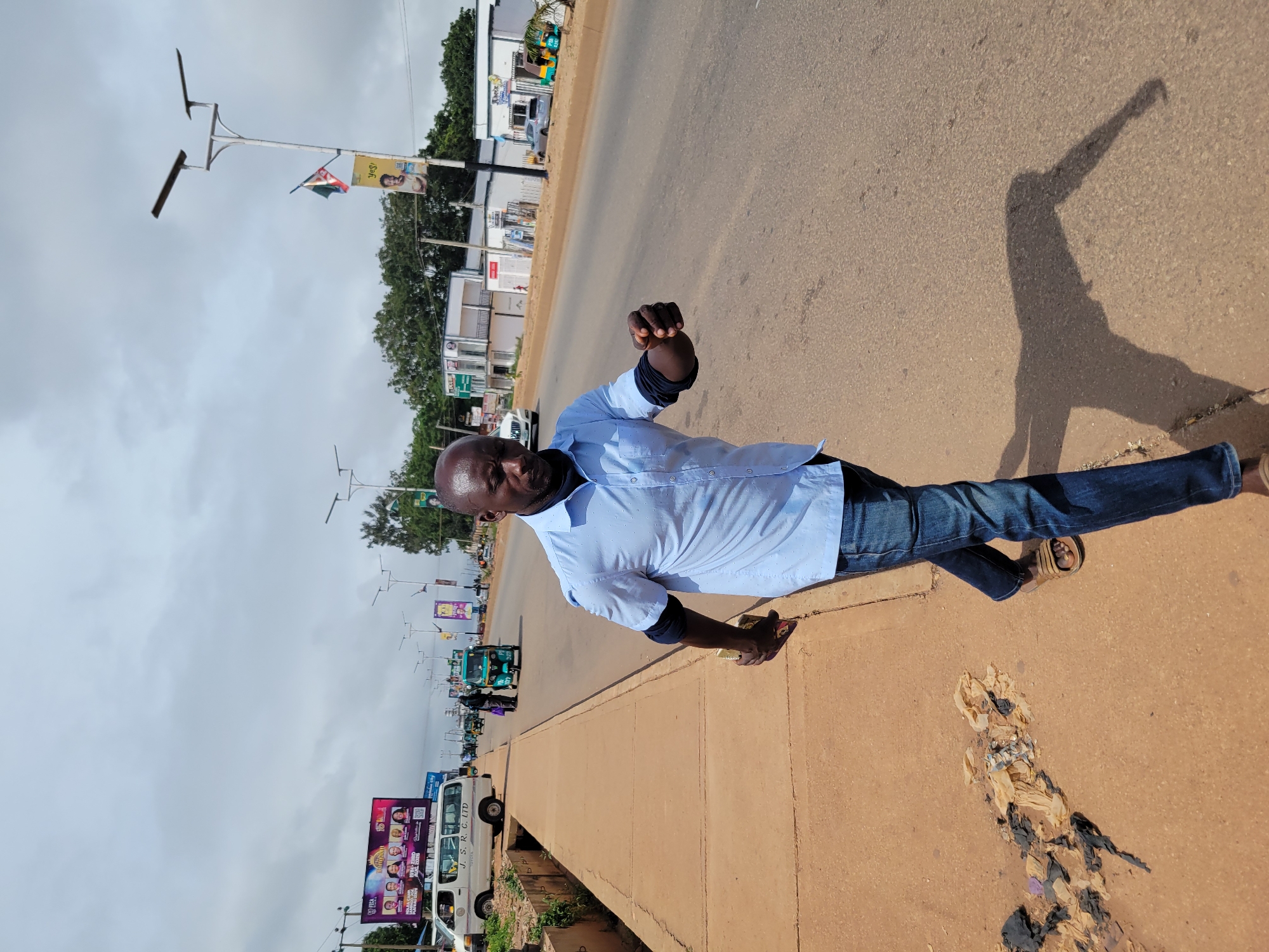 In Jos, Nigeria, a Black man wearing a light blue collared shirt, dark blue jeans, and brown sandals walks on a sidewalk next to a busy road. He is bald and has a dark complexion. His right arm is bent at the elbow, with his hand extended forward. He holds a yellow object in his left hand. Behind him, a white van with "J. S. R. C. LTD TOYOTA" written on the side is parked. Further down the road, several "Keke Napep" (three-wheeled taxis) and other vehicles are visible. Buildings line the opposite side of the road, with signs that are difficult to read. A large billboard displaying several people's faces is visible in the distance. The sky is overcast with clouds. The sidewalk is made of concrete and appears to have debris on it. The road is paved and appears to be in good condition.