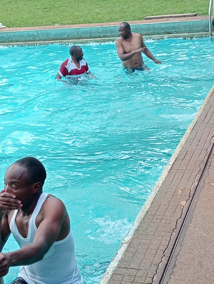 This media file captures three adult men enjoying a swimming pool at Riara Ridge, Kenya, during the daytime.
In the mid-ground, two men are standing chest-deep in the clear, blue water. The man on the left is wearing a red and white striped t-shirt and is looking towards the man on the right. The man on the right is shirtless, wearing dark swim trunks, and appears to be speaking or gesturing with his right hand as he looks at the other man.
In the foreground, partially cropped on the left, a third man is seen from the chest up, wearing a white tank top. He is holding onto the pool's edge, seemingly either entering or exiting the water. His left hand is covering his mouth, suggesting a reaction like surprise, amusement, or perhaps clearing his throat from water.
The setting is an outdoor swimming pool with light-colored concrete coping visible along the edge. Beyond the pool, a patch of vibrant green grass indicates a natural outdoor environment. The bright lighting suggests it is daytime. There is no visible text in the image.