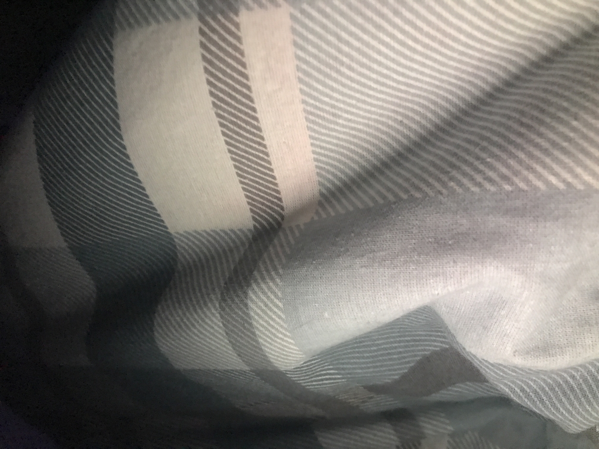 The image shows a close-up of a draped fabric with a gray and white plaid pattern. The fabric is bunched and folded, creating shadows and variations in color intensity. The pattern consists of alternating gray and white stripes, forming a grid-like design. There is no visible indication of a location in Yerevan, Armenia, nor any people, subjects, objects, actions, or interactions. The scene consists solely of the fabric itself.