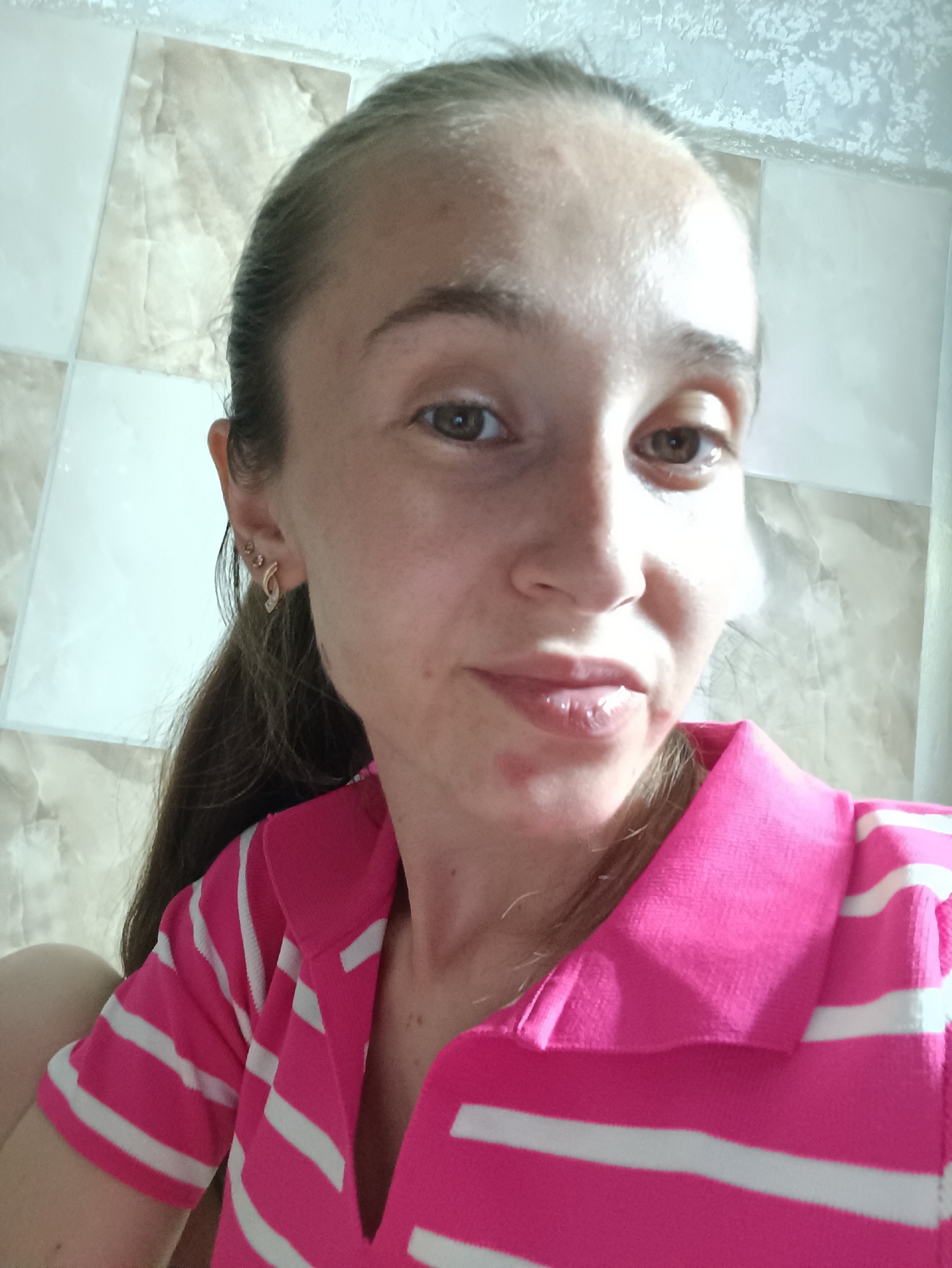 A woman with long brown hair pulled back is pictured from the chest up, looking at the camera. She is wearing a bright pink polo shirt with white horizontal stripes. The background appears to be a tiled wall with a marbled pattern. The woman has light skin and brown eyes, and she is wearing small earrings in her left ear. There are some red marks on her face around her mouth. The image appears to have been taken indoors. There is no visible indication of the location in the city of Vypasne, Ukraine.