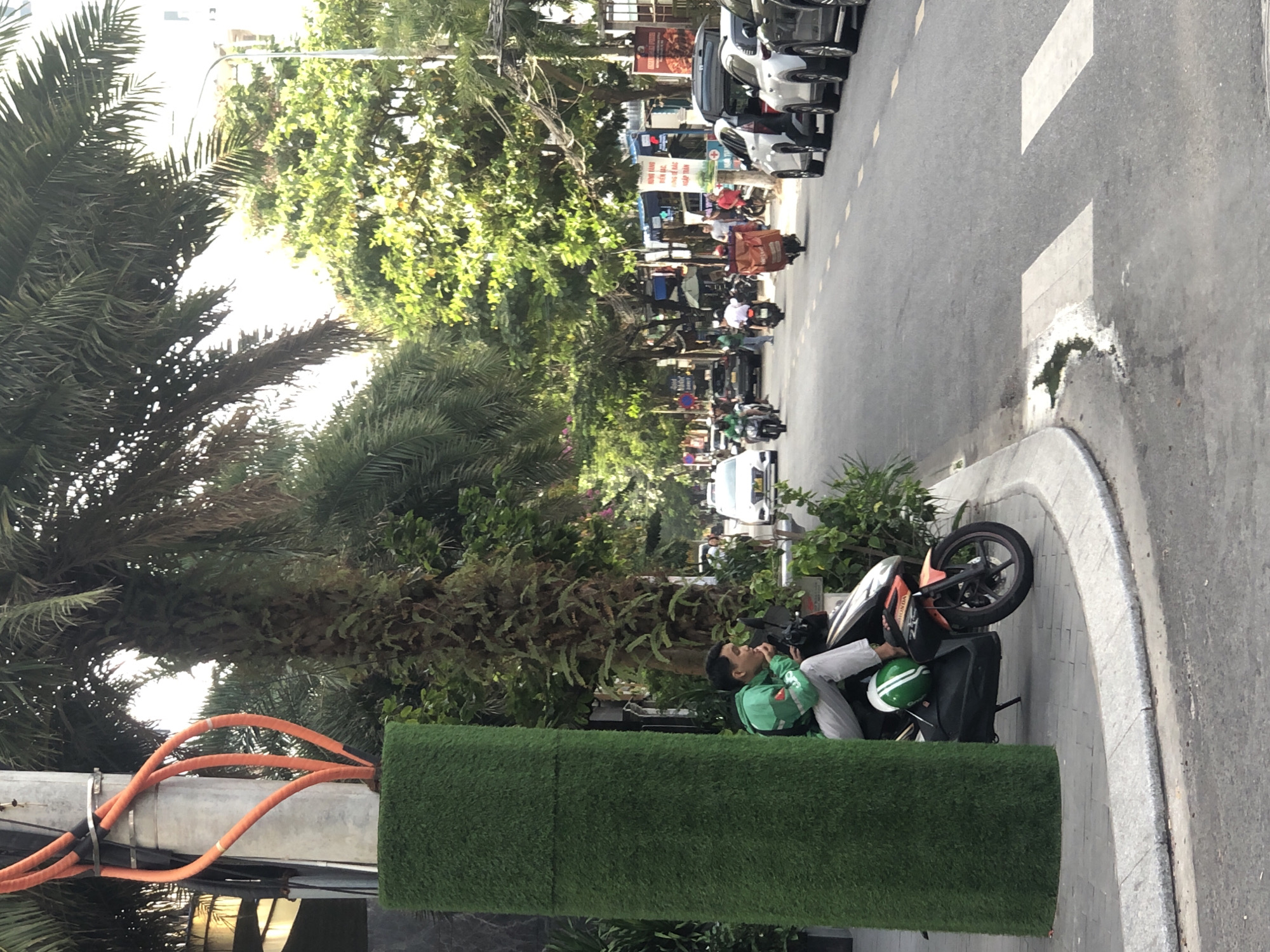 The image depicts a street scene in Đà Nẵng, Vietnam, with a focus on a man sitting on a scooter on the sidewalk. The man is wearing a green jacket and grey pants. A green and white helmet is placed on the scooter. The scooter is parked next to a large, cylindrical structure covered in artificial grass, which is next to a utility pole with orange cables. The street is lined with trees and buildings, and there is traffic on the road, including other scooters and cars. A pedestrian crossing is visible on the asphalt road. The overall atmosphere suggests a typical urban environment in Southeast Asia, with a blend of modern infrastructure and natural elements.