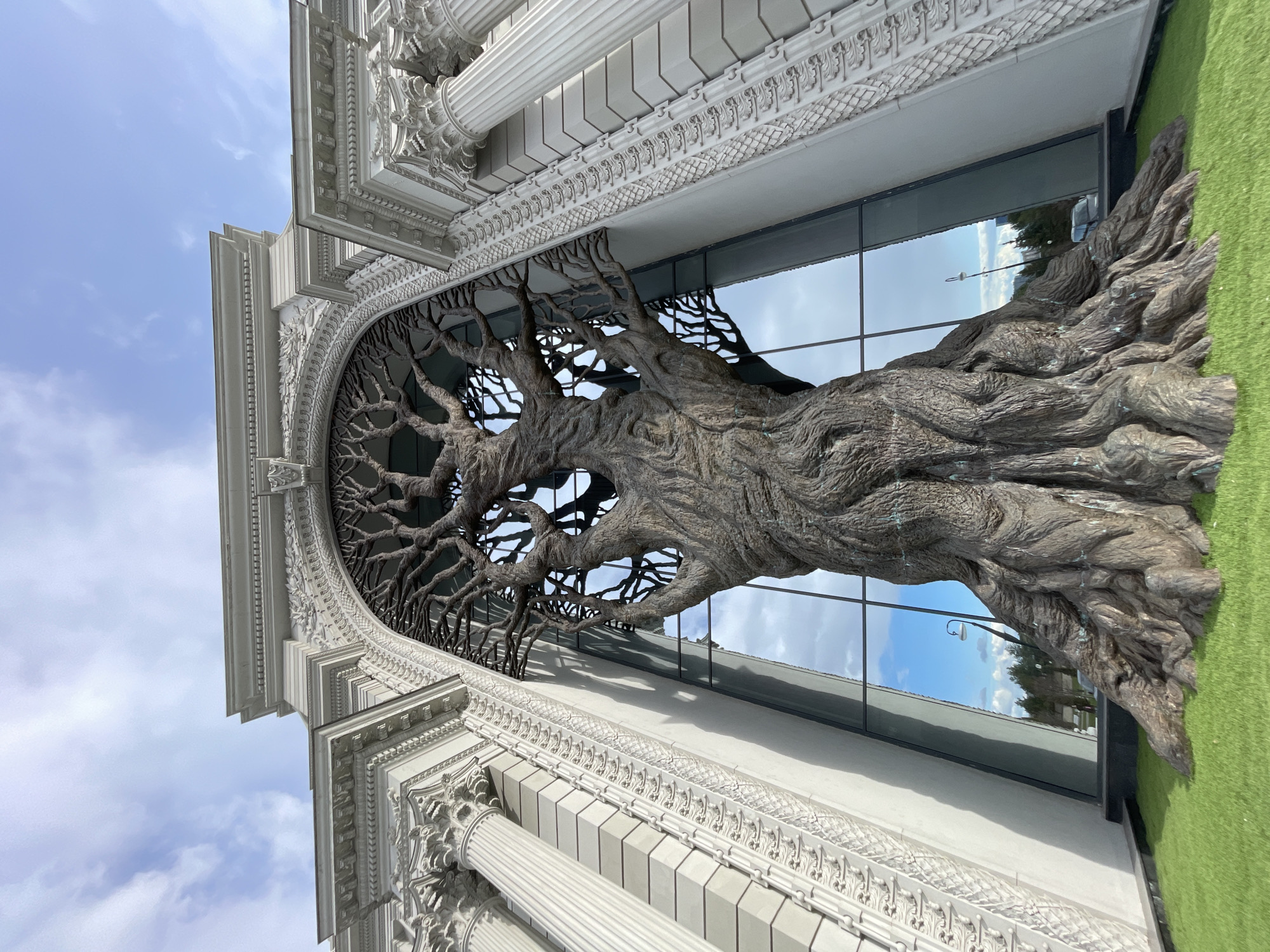 The image captures a grand architectural facade featuring a massive, intricately detailed tree sculpture. The sculpture, appearing to be made of a dark, textured material resembling weathered wood or bronze, forms a prominent artistic element across a large, arched window of the building. Its thick, gnarled trunk is visible at the bottom, with numerous branches spreading upwards and outwards to fill the archway, creating a complex, organic pattern against the glass.
The building itself is stately, constructed from a light-colored material, likely white stone or concrete. It exhibits classical architectural characteristics, including prominent fluted columns with ornate capitals, elaborate cornices, and intricately carved friezes featuring decorative patterns such as foliage and geometric motifs. These details suggest a building of significant cultural or public importance in Kazan, Russia.
Behind the tree sculpture, the expansive glass window reflects the bright blue sky dotted with scattered white clouds, indicating clear daytime weather. Reflections of distant trees, a streetlamp, and other parts of the urban landscape are also visible in the glass. A strip of vibrant green artificial turf lines the ground directly in front of the building and the base of the tree sculpture. No people are visible in the scene, and there is no discernible text. The overall impression is one of a striking artistic installation integrated with classical architecture under a bright sky.
