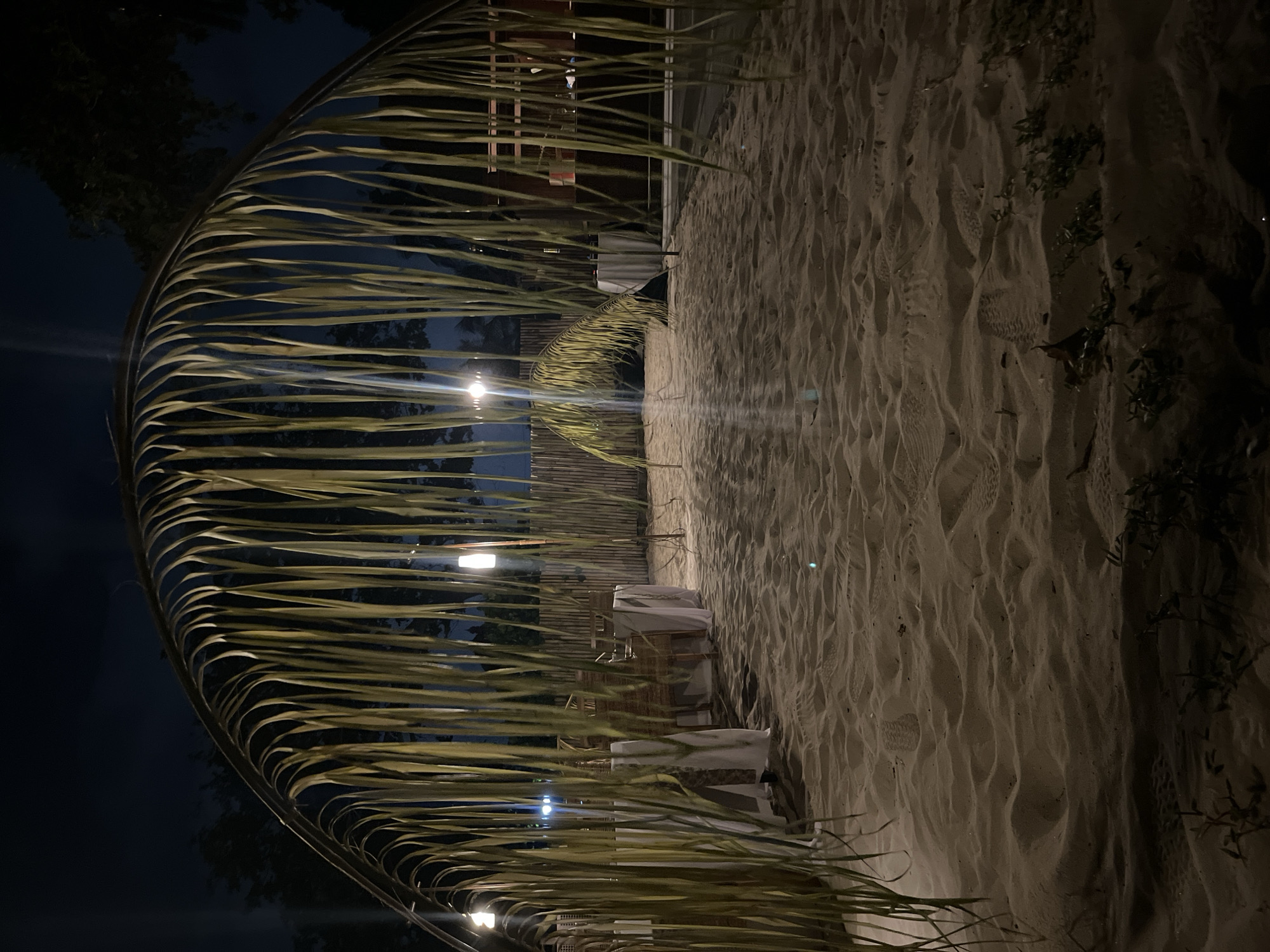 The image depicts a pathway on a sandy beach at night, likely in Kuta Selatan, Indonesia. The foreground is dominated by the textured sand, with some sparse vegetation at the bottom edge. A large, archway constructed from palm fronds forms a canopy over the pathway. Beyond the archway, several tables draped with white tablecloths are visible, suggesting a dining or event setup. These tables are accompanied by chairs, some of which appear to be made of woven material. A bamboo fence lines the background, and lights illuminate the scene from behind the fence and within the dining area. The overall atmosphere is dark, with artificial lighting creating pools of illumination on the sand and around the tables. There are no people visible in the image.