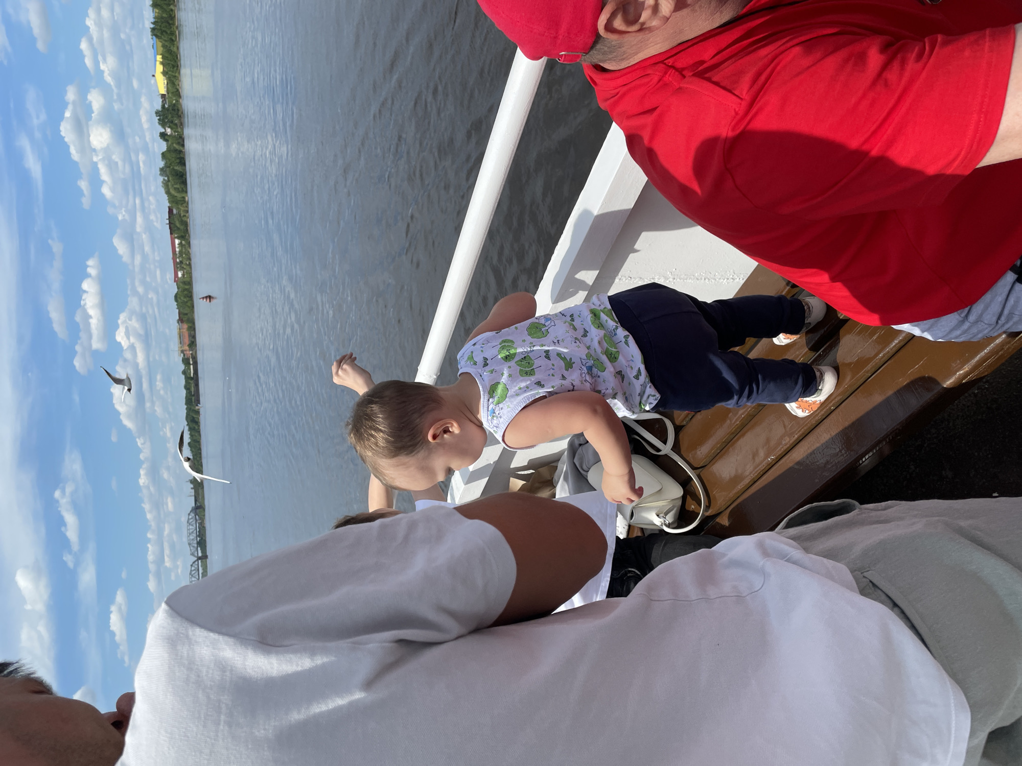 This image depicts a bright, sunny daytime scene aboard a boat or ferry, likely on the Kama River in Perm, Russia. The main subject is a young child, appearing to be a toddler, dressed in a white top with a green pattern and dark trousers, leaning over a white railing to look out at the water.
Several adults are partially visible around the child. On the right, an adult in a red shirt and red cap is positioned behind the child. In the foreground, another adult wearing a white short-sleeved shirt is seen from the side. A white handbag rests on a brown wooden bench or deck beside the child, with a grey garment (possibly a hoodie) also present.
The sky is clear blue with scattered white clouds, indicating pleasant weather. The expansive body of water, characteristic of a large river, stretches into the distance. On the far bank, a tree-lined shore is visible, dotted with buildings, some with yellow or red roofs. A prominent bridge structure can be seen in the distance, spanning the river. Two seagulls are captured mid-flight against the blue sky. A red buoy is also visible in the calm river water. The scene conveys a sense of a relaxed outing or journey on the water. No visible text is discernable in the image.