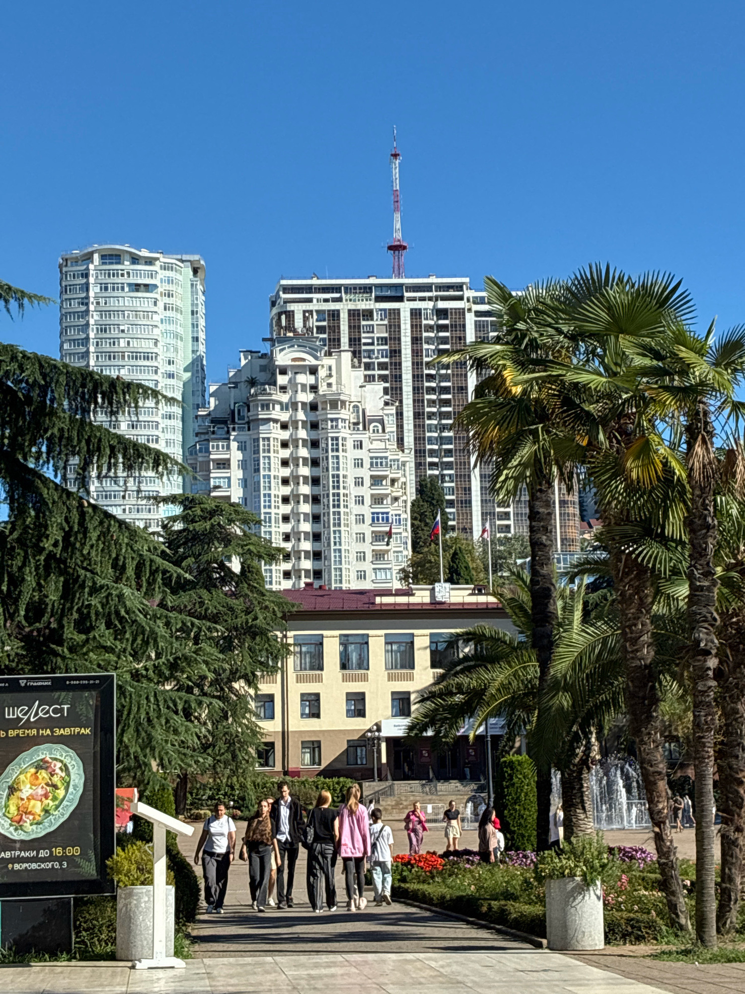 The image captures a sunny day scene in the city of Sochi, Russia, with a prominent television tower and several modern high-rise buildings dominating the skyline. In the foreground, lush green trees and palm trees frame the view. A group of people are walking along a paved path, some are dressed casually, others are in more formal attire. One person appears to be looking at a large advertisement billboard. Behind them, a fountain can be seen in the distance, and several cars are parked on a street. The overall atmosphere is one of a bustling urban environment with elements of a resort city.