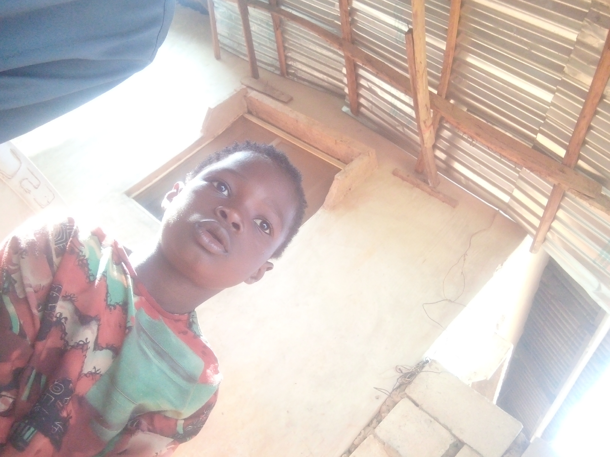 A photograph, undated and unverified, shows an upward-angled view featuring a child in the lower left quadrant of the frame, with a backdrop depicting a building's interior. The child is dark-skinned, appearing to gaze upwards, wearing a multi-colored shirt. Above the child, a portion of a person's leg, clad in blue fabric, intrudes into the upper left corner. The background includes a section of a wall, a window opening, and a corrugated metal roof supported by wooden beams. The location is noted as Unknown, Unknown. The image's lighting appears to be bright, potentially from daylight streaming through the openings.