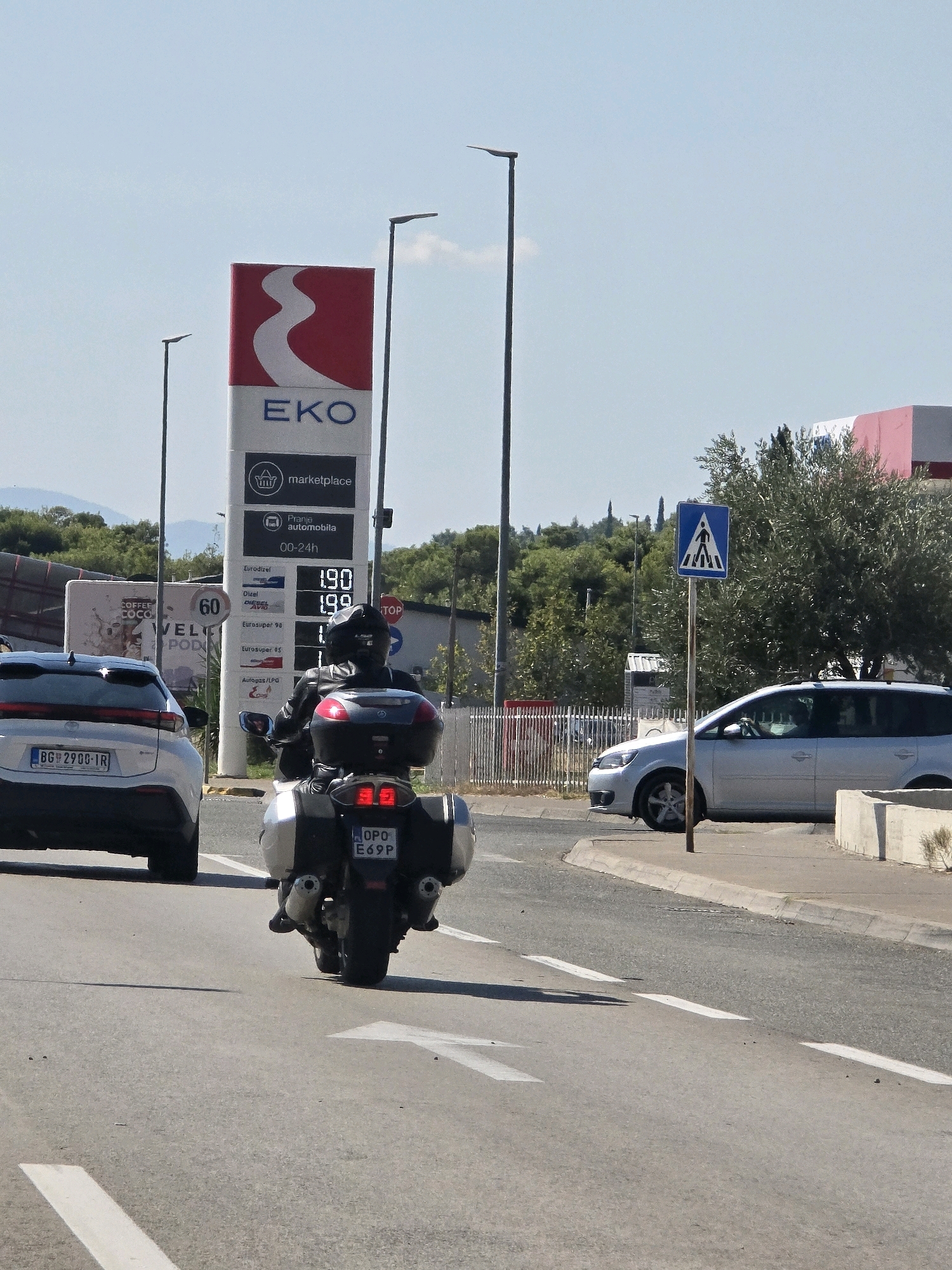 An outdoor, eye-level, medium shot captures a sunny daytime scene on a road in Podgorica, Montenegro. In the foreground, a person rides a black and silver motorcycle with saddlebags, heading away from the camera. The motorcycle has a license plate reading "OPO E69P". To the left of the motorcycle, a white car with a license plate reading "BG 2908-1R" is visible, also moving away from the viewer.
Dominating the mid-ground on the right is a tall gas station sign for "EKO" and "marketplace", displaying fuel prices of 1.90 and 1.99 for "Eurodizel" and "Diesel Avio" respectively, and 1.88 for "Eurosuper 98". The sign indicates "Pranje automobila" (Car wash) and operating hours "00-24h". A pedestrian crossing sign with a blue background and a white symbol of a person walking is positioned to the right of the gas station sign, near a silver station wagon.
In the background, lush green trees and hills under a clear blue sky suggest a location with natural beauty. The road surface is asphalt with white lane markings. The overall impression is of a typical roadside scene with vehicles and essential services in a bright, urban environment.