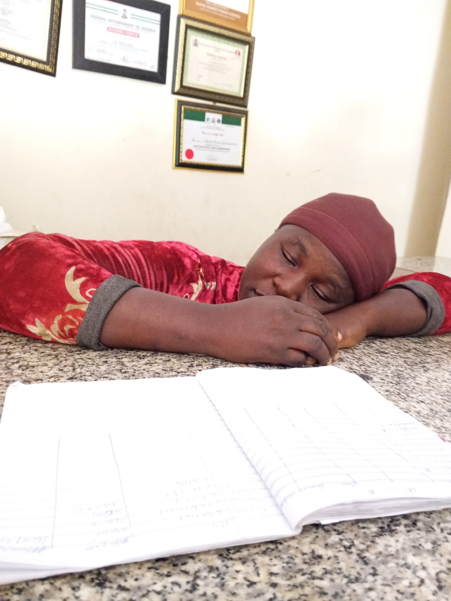 The image captures a person sleeping or resting with their head on a speckled table, supported by their folded arms. The individual, who appears to be an adult, is wearing a deep red, possibly velvet, long-sleeved top adorned with gold patterns, and a dark reddish-brown head covering. Their eyes are closed.
An open, lined notebook with handwritten text and vertical columns lies on the table in front of the sleeping person. The text in the notebook is illegible.
The background reveals a light-colored wall decorated with several framed certificates or official documents. One certificate, prominently displayed in a black frame, clearly shows "FEDERAL GOVERNMENT OF NIGERIA" at the top. Another certificate, positioned lower and to its right, features a green and white logo, possibly "E.A.O," and a red circular seal. The text on these background documents is mostly unreadable due to focus and distance.
The setting is indoors, possibly an office, administrative area, or study environment, suggested by the presence of framed certificates. The lighting is uniform, indicating an indoor scene. No specific time of day or weather conditions can be determined from the photograph. The atmosphere is calm and quiet, emphasizing the person's state of rest.