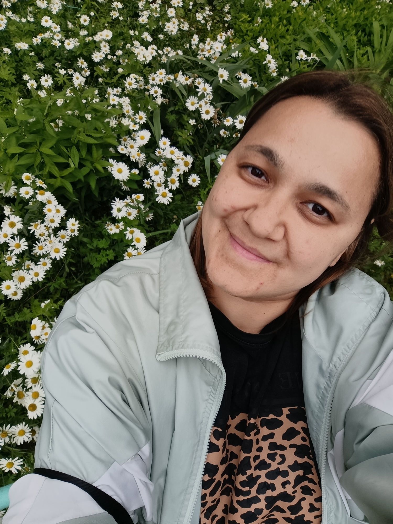 A selfie is taken by a person sitting in a field of white daisies. The person has brown hair and is smiling, looking towards the camera. They are wearing a light blue jacket over a black t-shirt with a leopard print design. The background is filled with numerous small white daisies and green foliage. The lighting suggests it is daytime, and the scene appears to be outdoors in a natural setting. No visible text is present in the image. The setting is implied to be Chaikovsky, Russia.