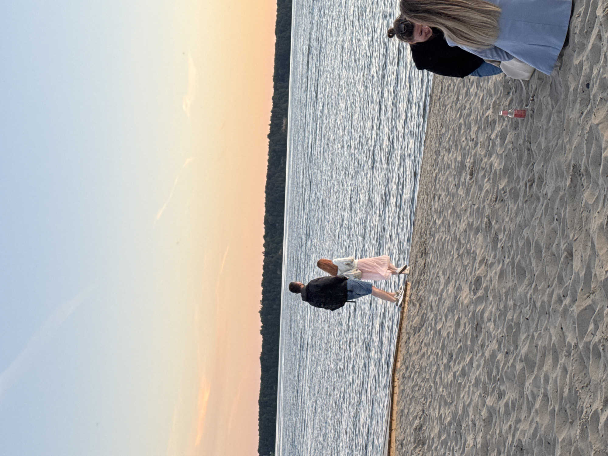 The image shows a beach scene with people and a body of water under a sunset sky. In the foreground, two women are on the sand; one is wearing a black hooded jacket and blue jeans, and the other is wearing a light blue jacket. A bottle of soda is on the sand between them. In the middle ground, a couple walks along the water's edge on a wooden boardwalk. The man is wearing a black backpack and shorts, and the woman is wearing a pink skirt and white jacket. The background features a tree-lined horizon and a calm body of water. The sky displays a gradient of light blue to orange, indicative of a sunset. The location is identified as a beach in the city of Tallinn, Estonia.