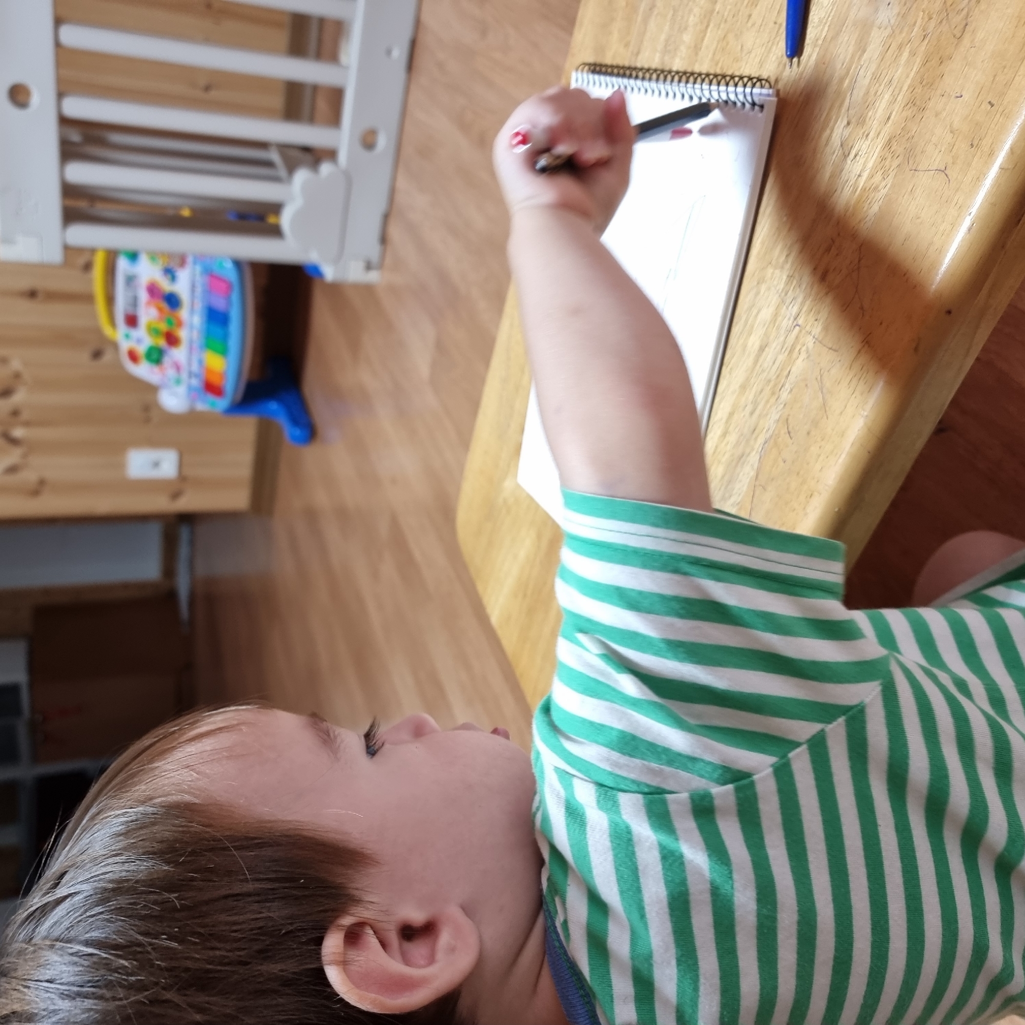 A young child, facing right in profile, sits at a wooden table, drawing in a notebook with a pen. The child has brown hair and wears a green and white striped t-shirt. The notebook is open to a page with faint lines, and the pen appears to be red. A blue pen lies on the table to the right. In the background, out of focus, is a white baby crib and a colorful toy keyboard. The flooring is wood. The scene is indoors, likely in a home.
