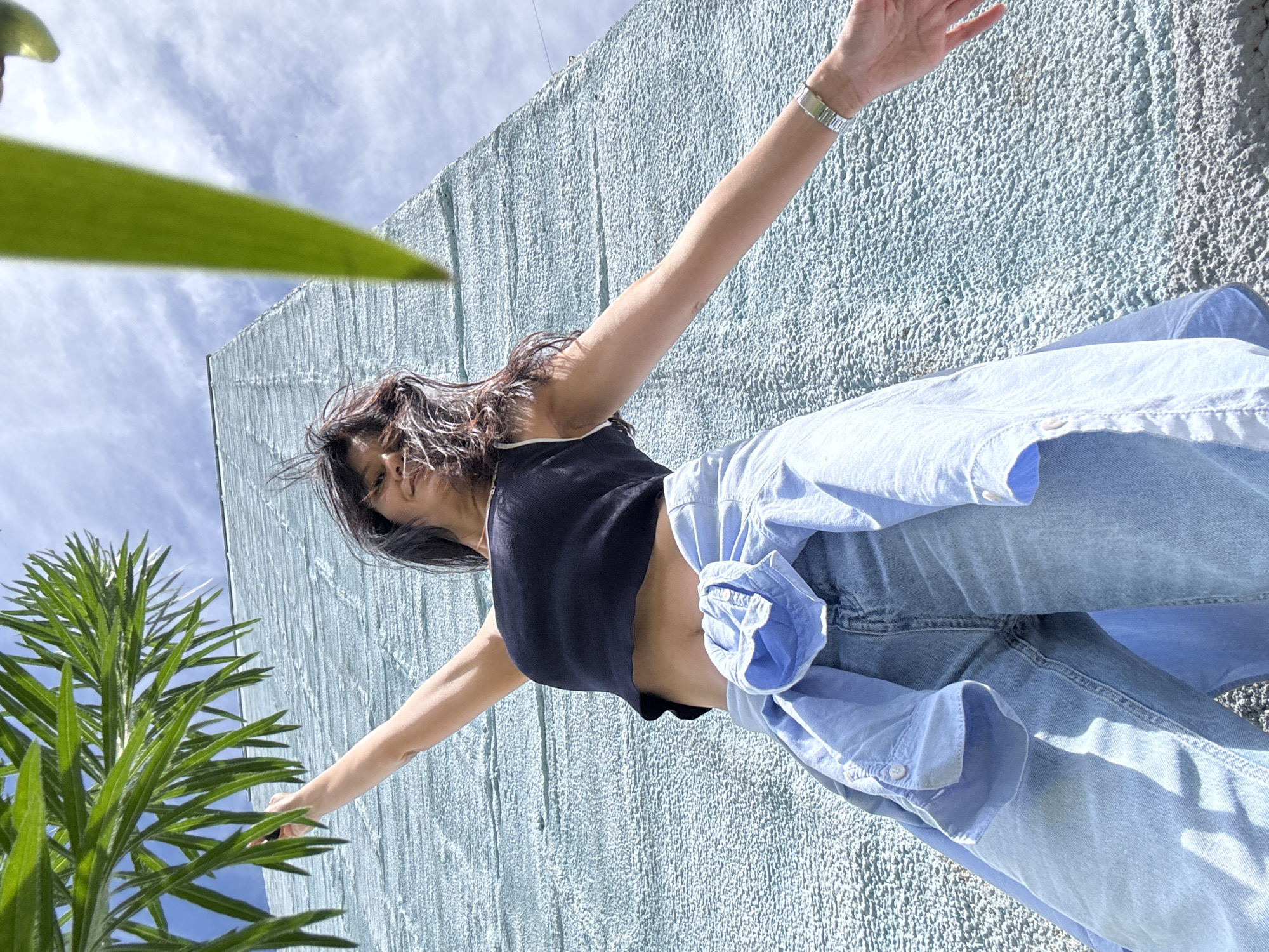 A low-angle, full-shot photograph shows a young woman standing in front of a textured, light blue wall in Chaikovsky, Russia. She is wearing a black crop top, a light blue unbuttoned shirt tied around her waist, and light wash jeans. Her arms are outstretched, and her hair is windswept. Green foliage is visible in the foreground, framing the upper left and right corners of the image, and the sky is partly cloudy. The texture of the wall suggests it might be stucco or a similar material.