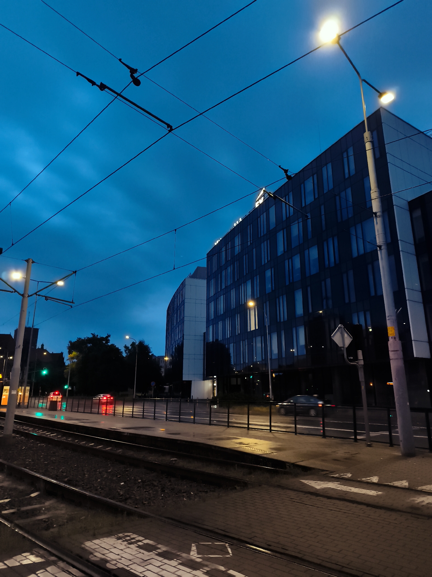 The image depicts a twilight street scene in Gdańsk, Poland, focusing on the area near the Olivia Star building. The sky is a deep, vibrant blue, suggesting either early evening or late dusk. Prominently featured are overhead tram wires and a streetlamp casting a warm glow. In the background, a modern, multi-story building with dark, reflective glass windows dominates the skyline. To the left, the tram tracks run parallel to the sidewalk, with a crosswalk marked with white arrows on the pavement. Several cars are visible on the wet street, their headlights and taillights creating streaks of light. A traffic light displays a green signal. The overall atmosphere is one of urban activity transitioning into night, with elements of public transportation and modern architecture characteristic of a developed city.